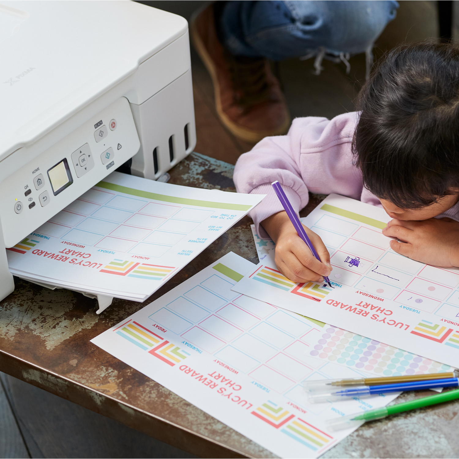 Dziecko rysuje na tabeli nagród fioletowym długopisem. Drukarka jest w tle.