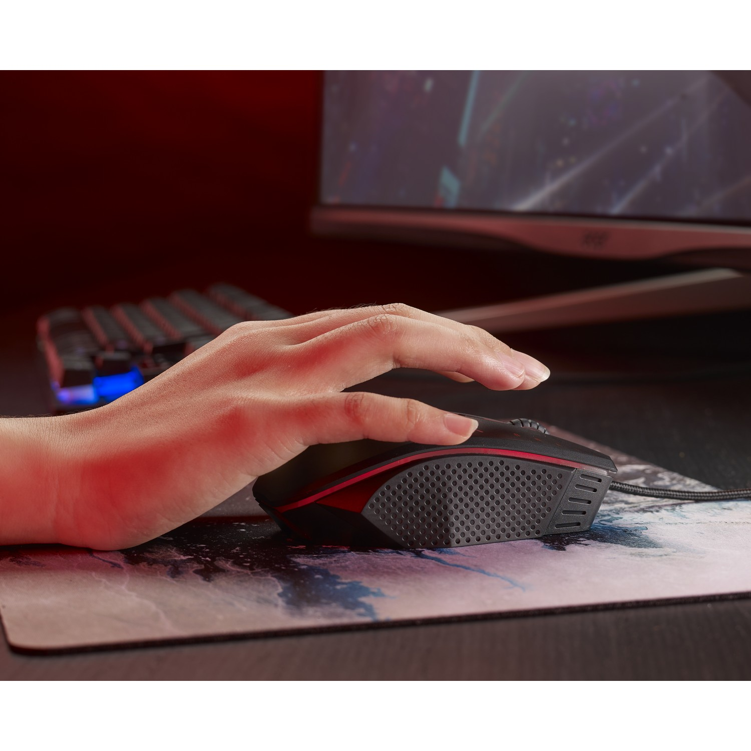 Mano su un mouse da gioco. Tastiera e monitor sullo sfondo. Il tappetino ha un design ad acquerello.