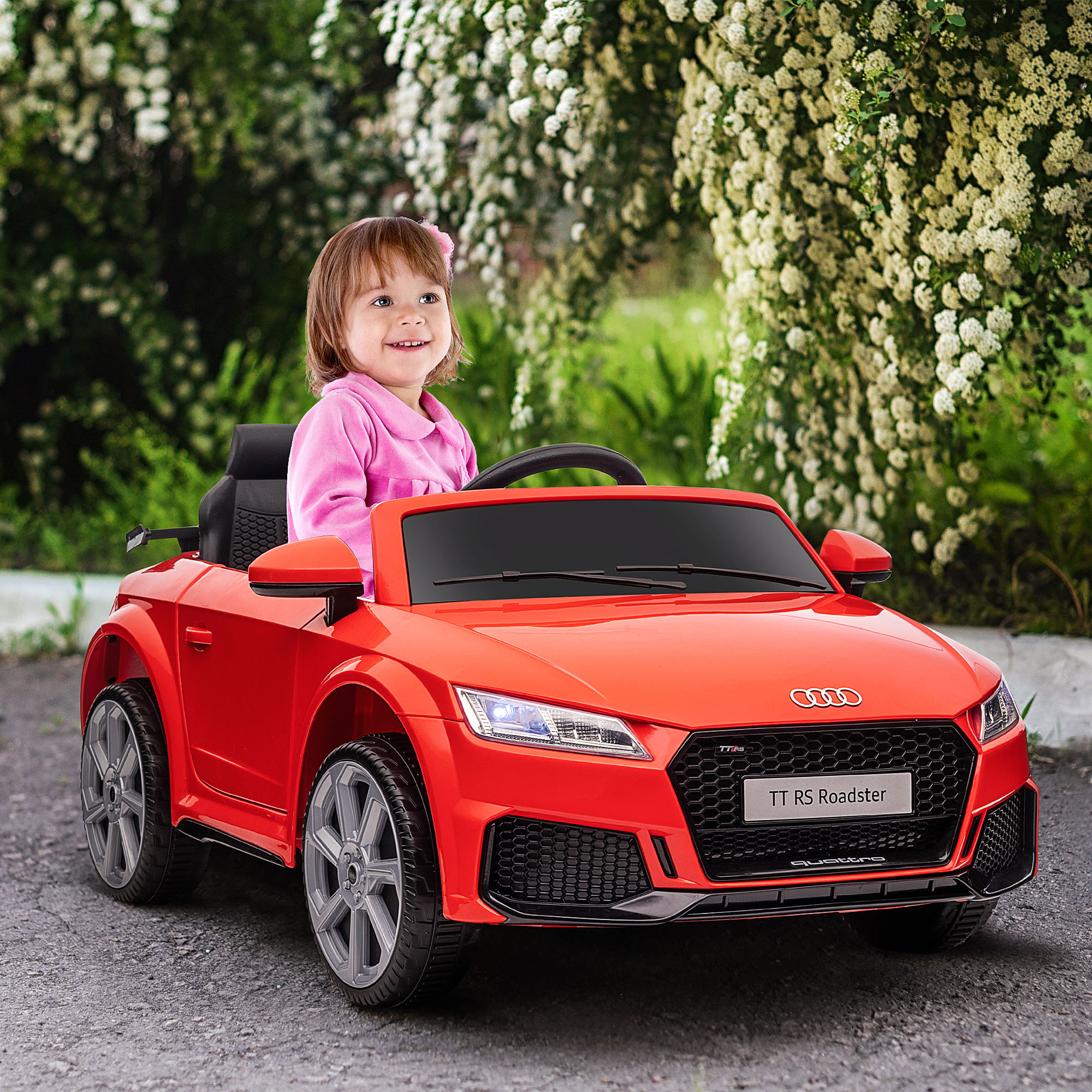 Una niña sonriente en un coche de juguete rojo. Flores blancas y hojas verdes al fondo.