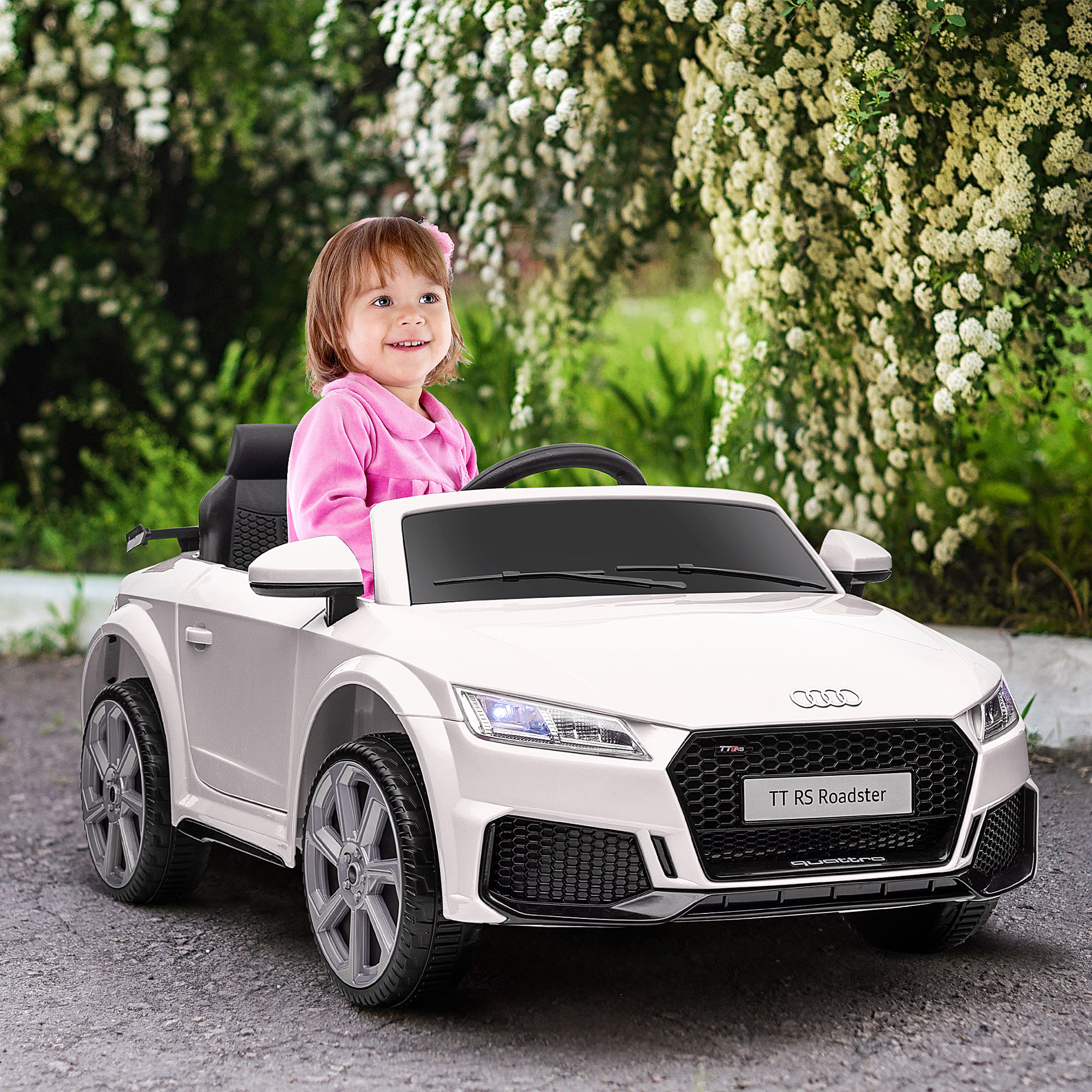 Una ragazzina con un top rosa guida un'auto giocattolo Audi TT RS Roadster bianca su una strada.