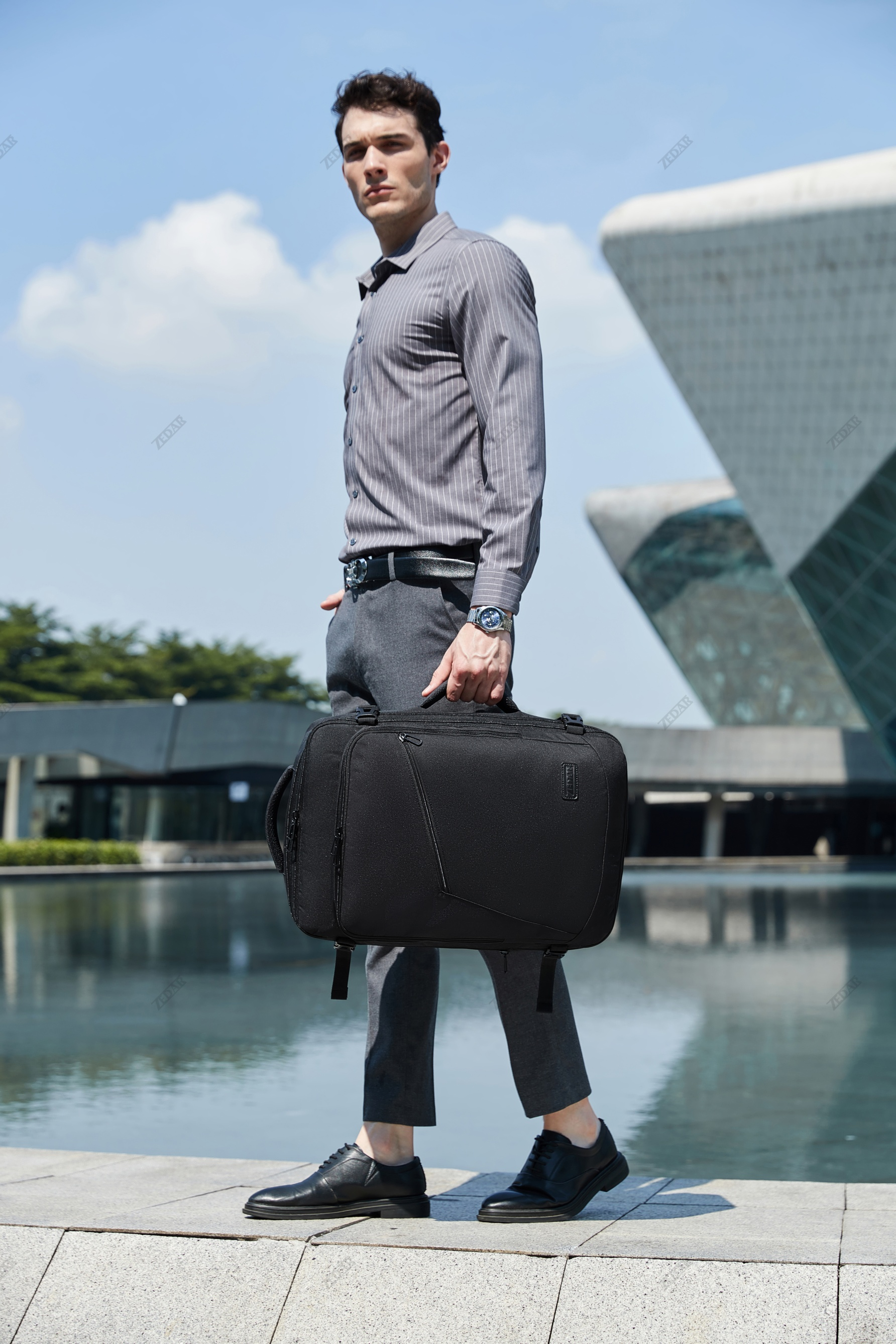 A man in a gray shirt carrying a black bag with a lake and building in the background.