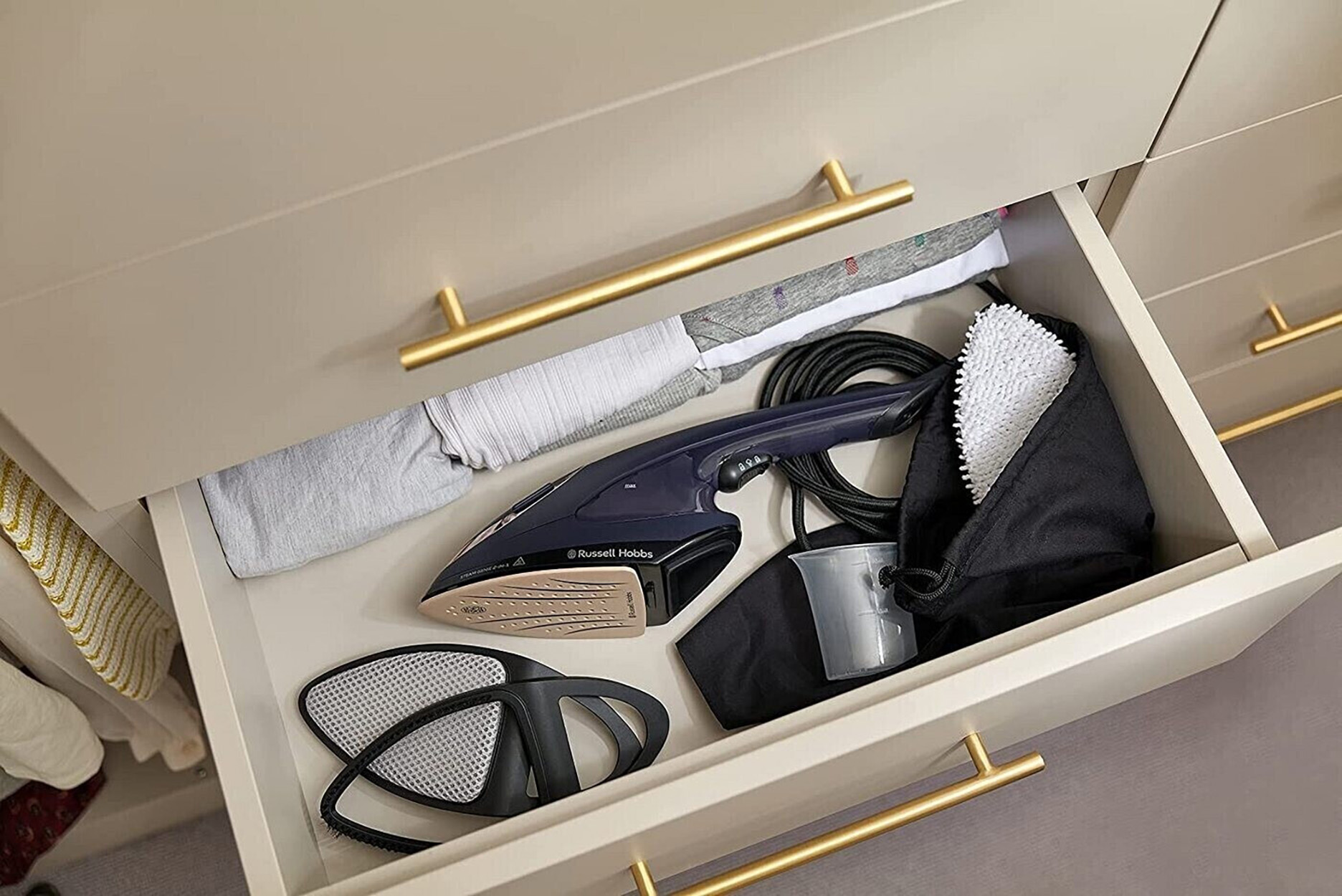 Otwarta szuflada z żelazkiem, ubraniami i akcesoriami. Złote uchwyty. Beżowa szafka.