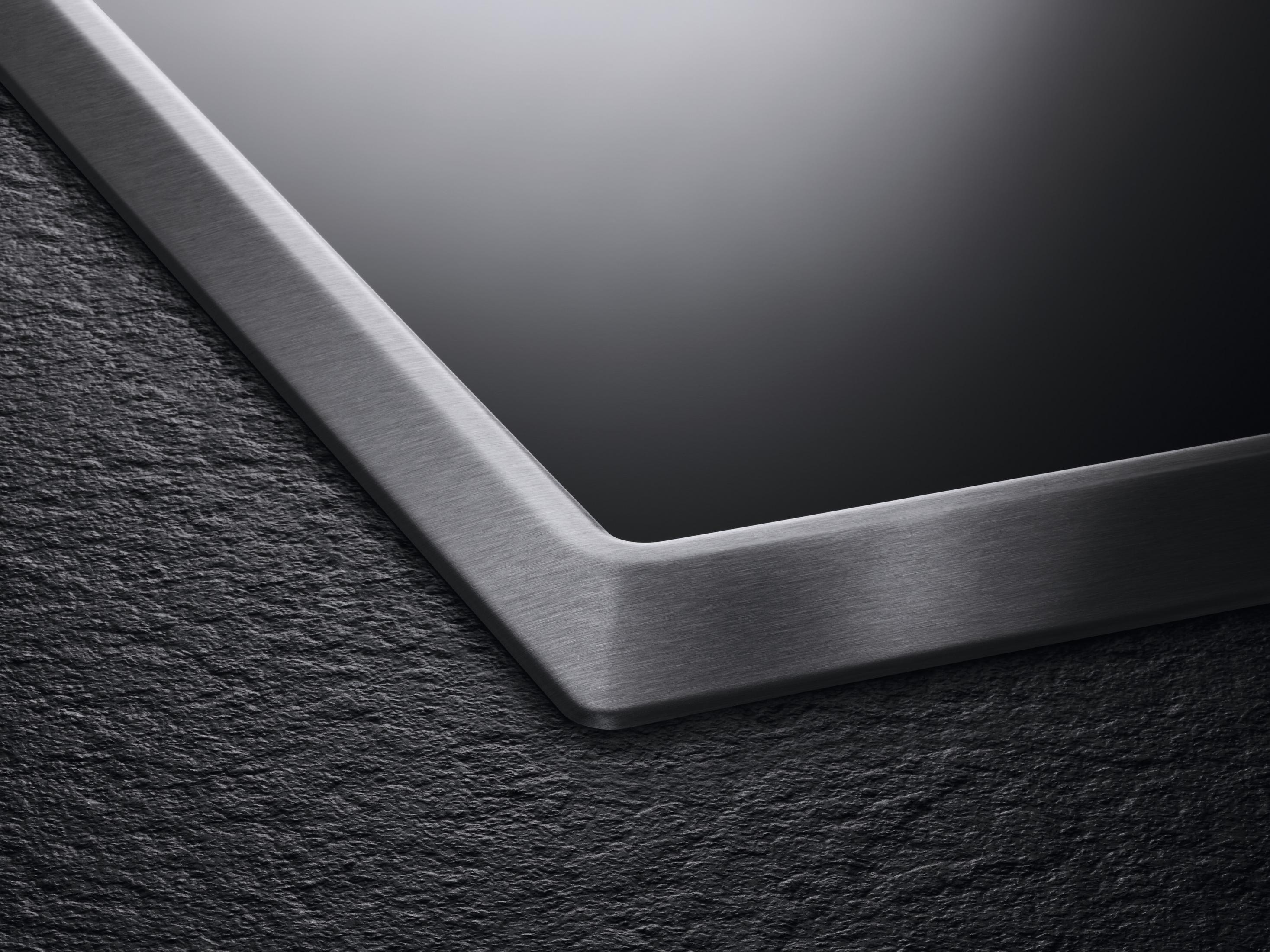 Zbliżenie na krawędź czarnej płyty grzewczej ze szklaną powierzchnią i ramą ze stali nierdzewnej, na ciemnej kamiennej powierzchni.