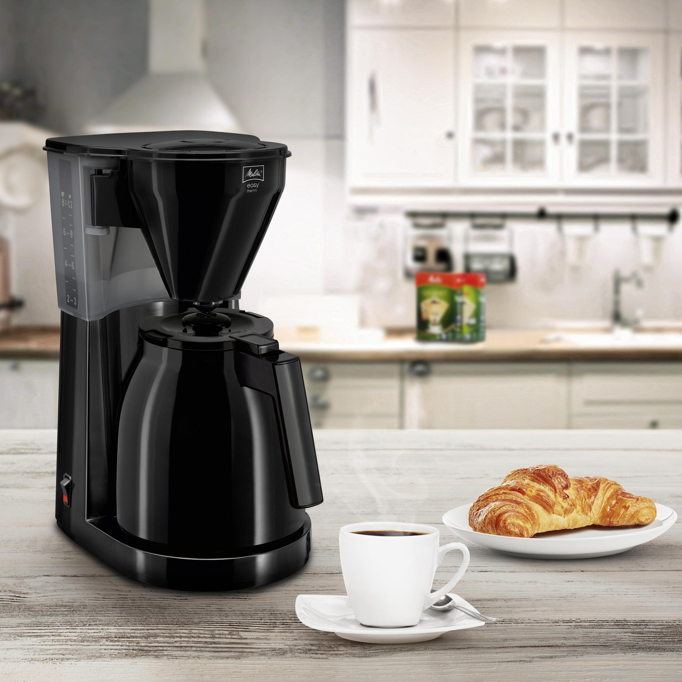 Czarny ekspres do kawy, filiżanka kawy i rogalik na stole.