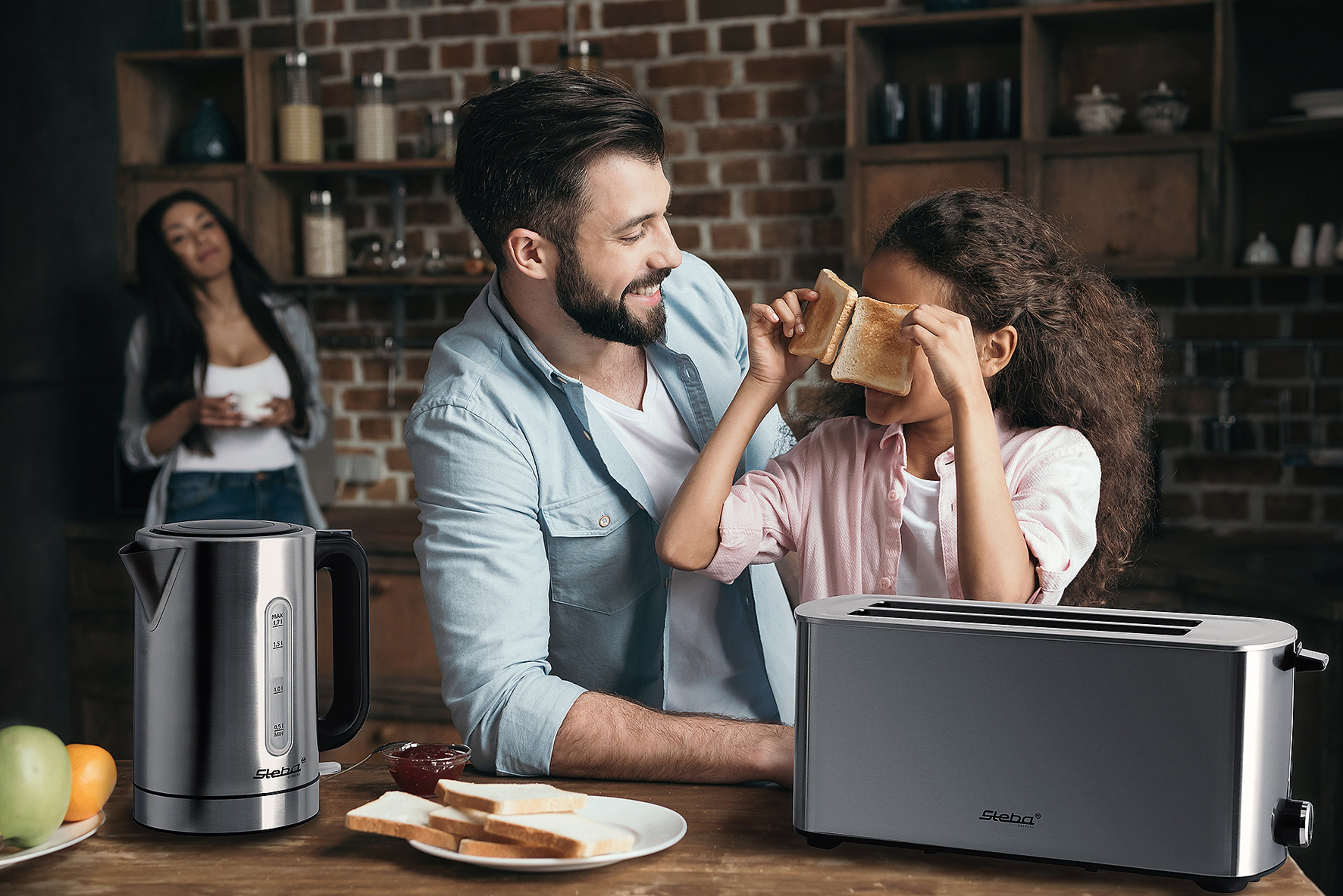 Familie in einer Küche. Vater und Tochter lächeln, die Tochter hält Toast. Wasserkocher und Toaster auf der Theke.