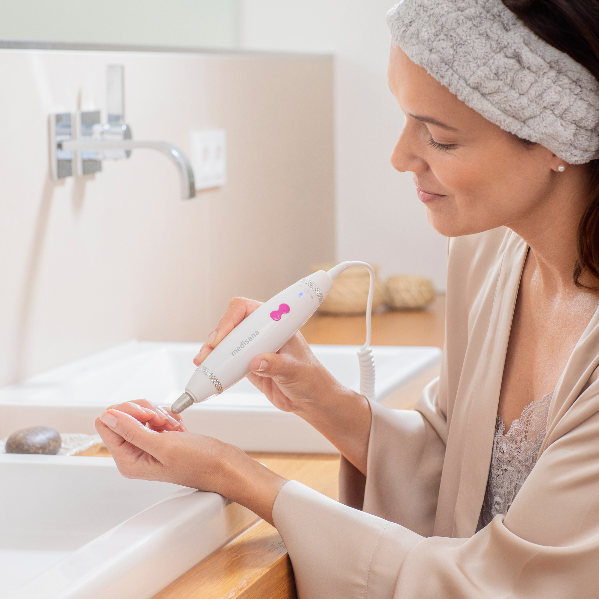 Frau benutzt ein weißes Nagelpflegegerät. Badezimmer mit Waschbecken im Hintergrund.