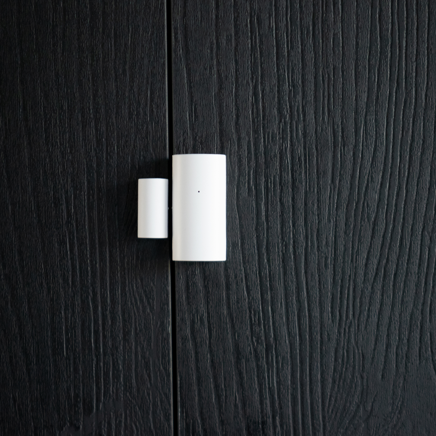 Weißer Smart-Home-Sensor an einer schwarzen Holztür. Er hat zwei Teile. Dunkler Hintergrund.