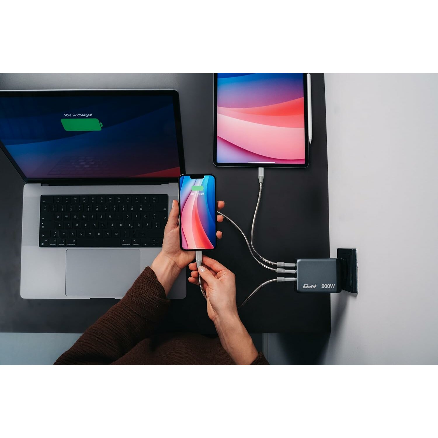 Kişi telefonu şarj cihazına bağlıyor. Yakınlarda dizüstü bilgisayar ve tablet. Görünümde güç hub'ı.