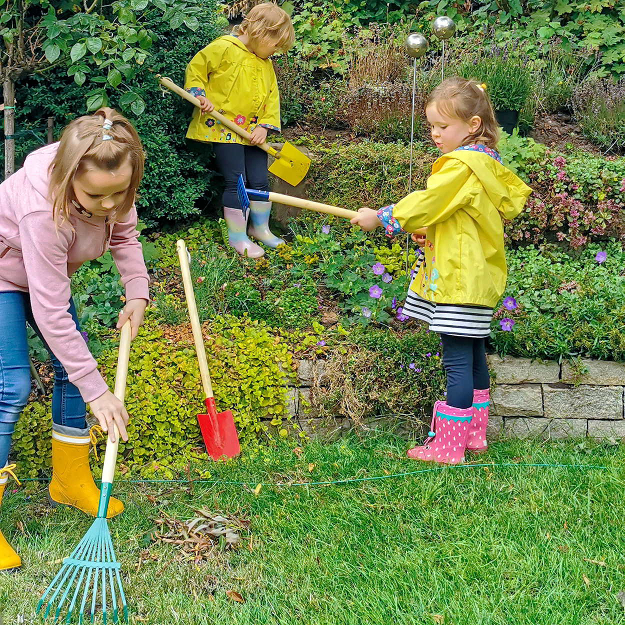 Tre bambini stanno giardinando in un giardino. Uno rastrella, uno zappa e uno usa un rastrello.