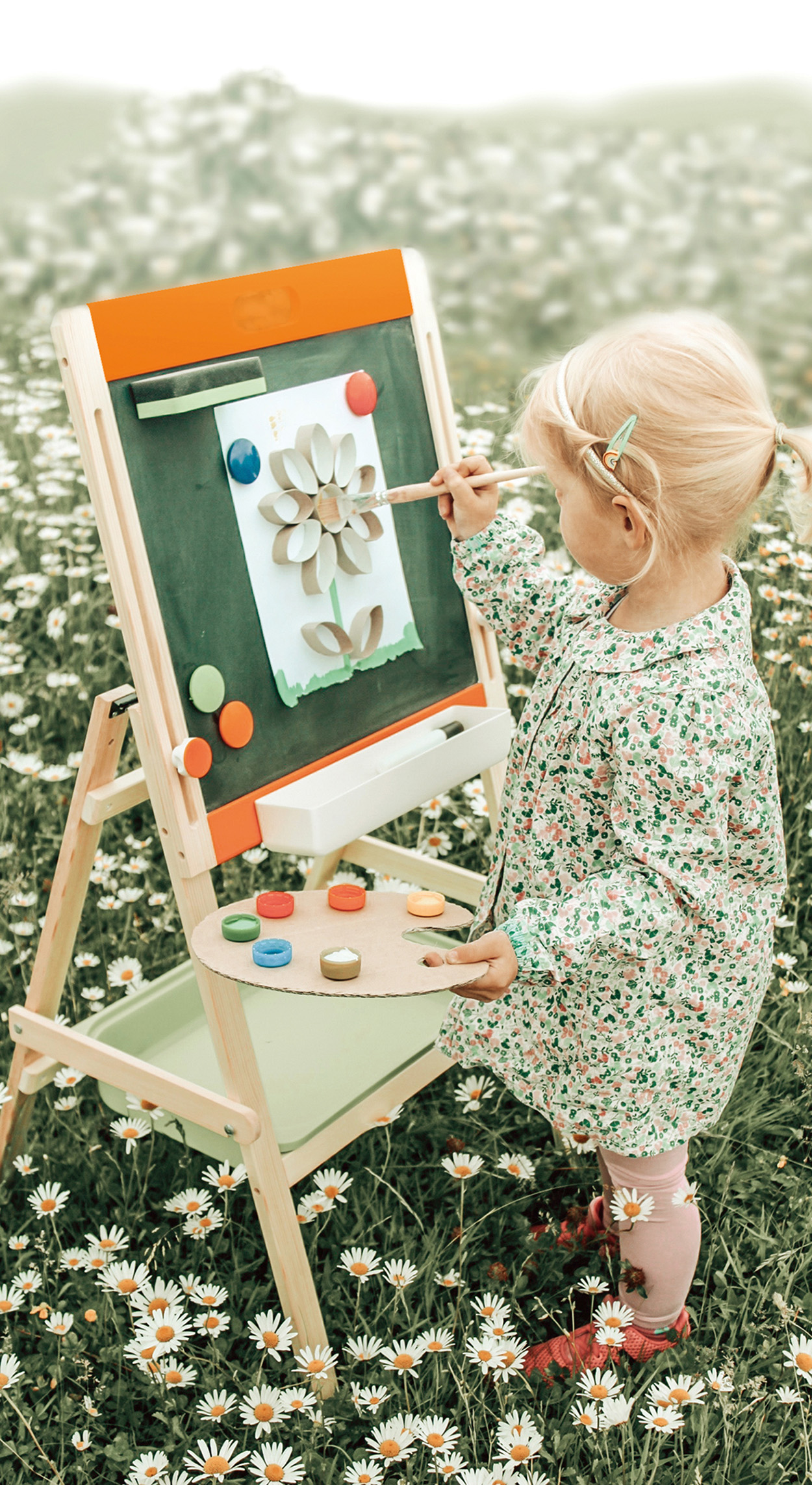 Kind malt auf einer Staffelei in einem Blumenfeld. Die Staffelei zeigt eine Papierblume.