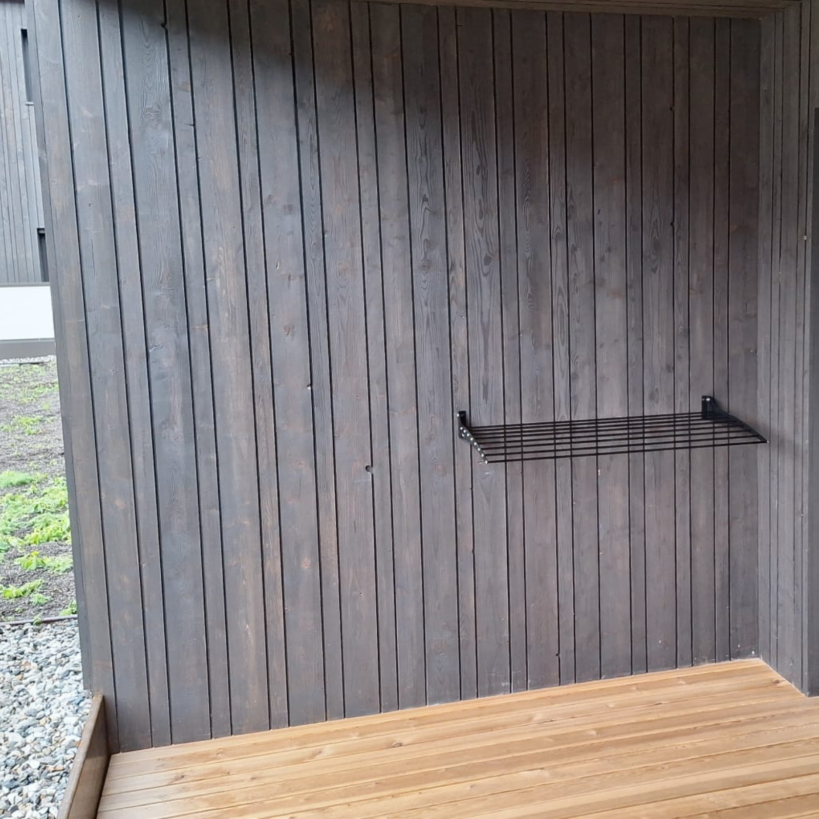 Uno stendibiancheria nero a parete su un ponte di legno. Il muro è marrone scuro, a doghe verticali.
