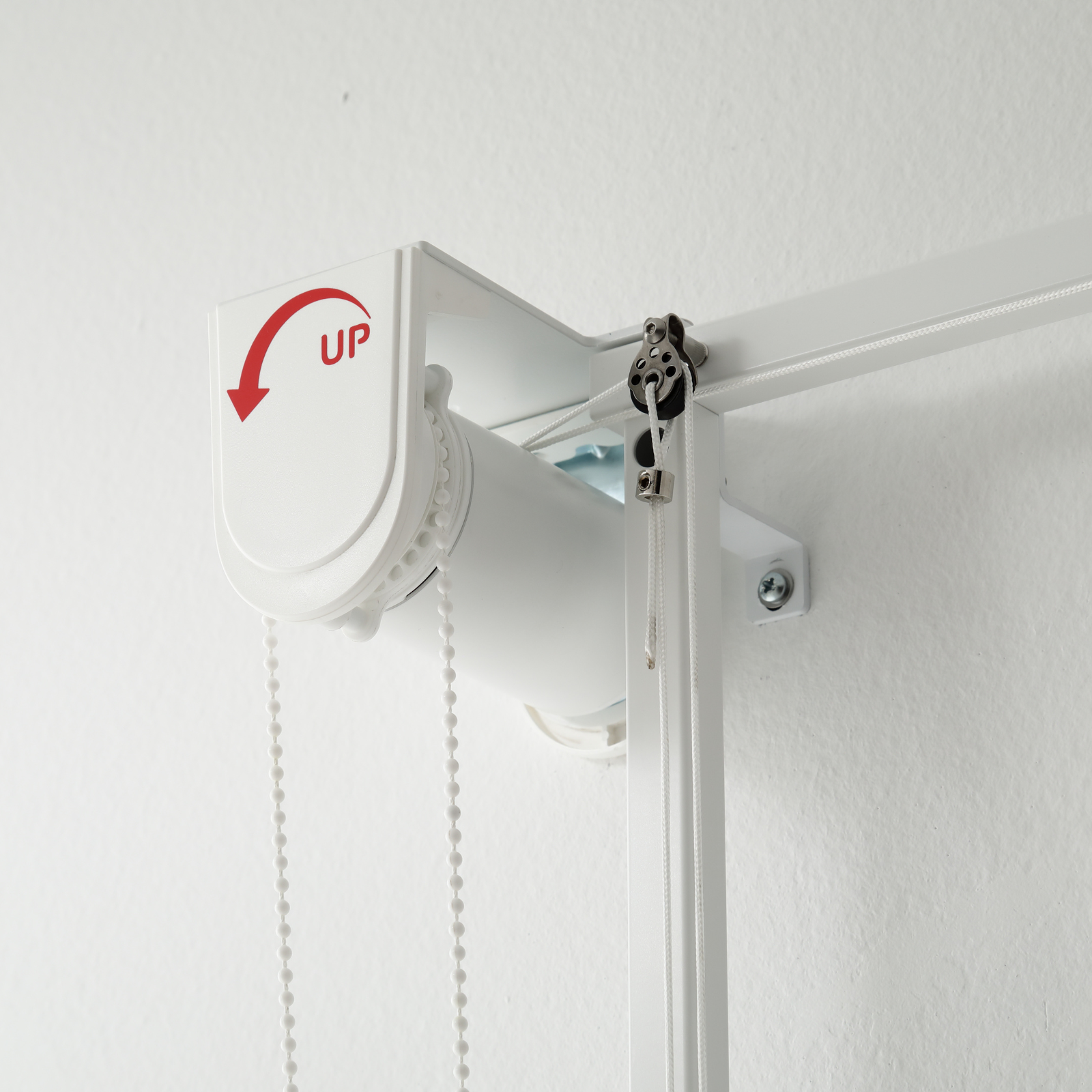 Un meccanismo per tenda a rullo bianco su un muro bianco, con freccia 'UP' e una catena.