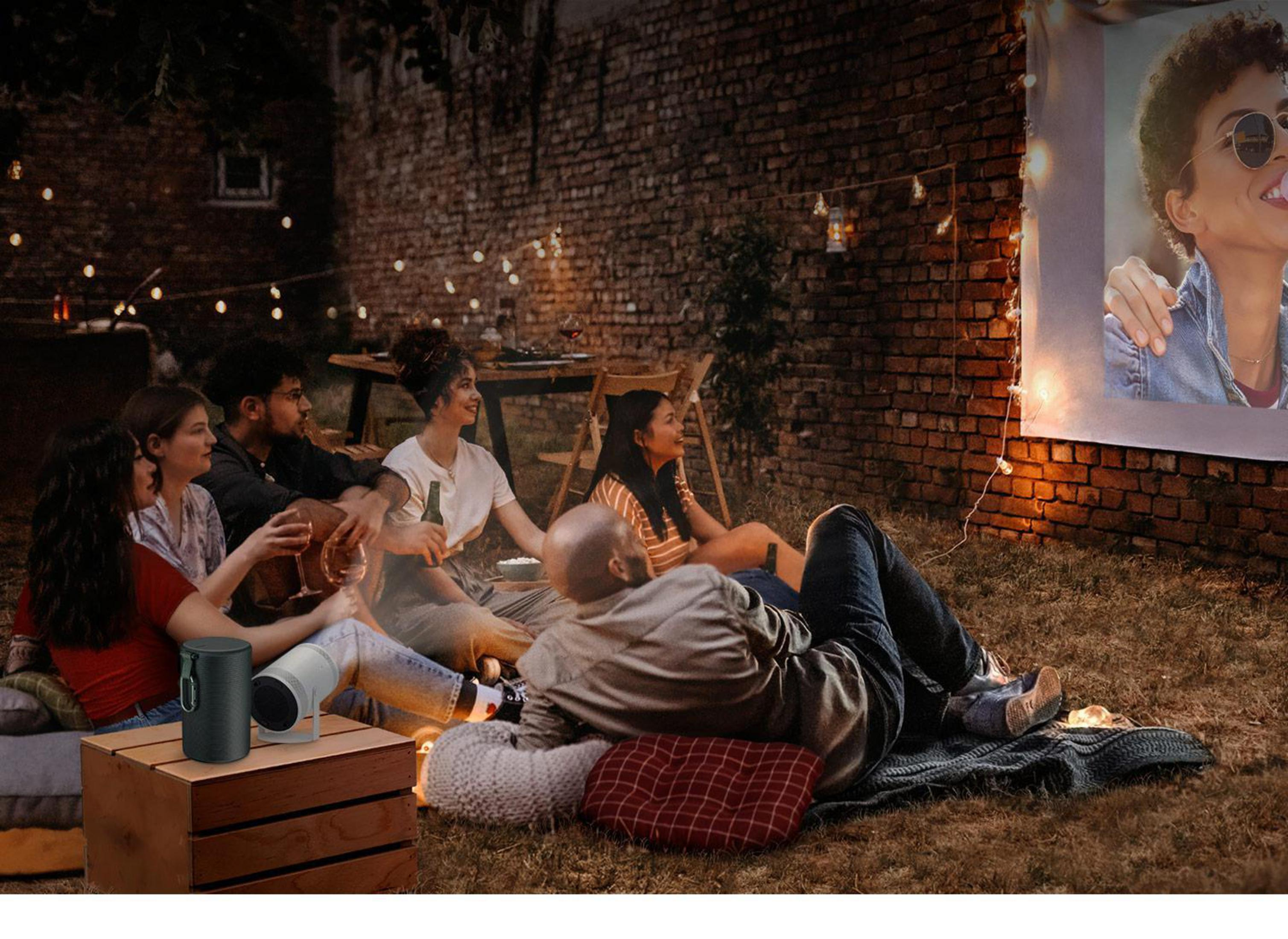 Grupo de personas viendo una película en una pantalla al aire libre. Proyector y altavoces en una caja de madera.