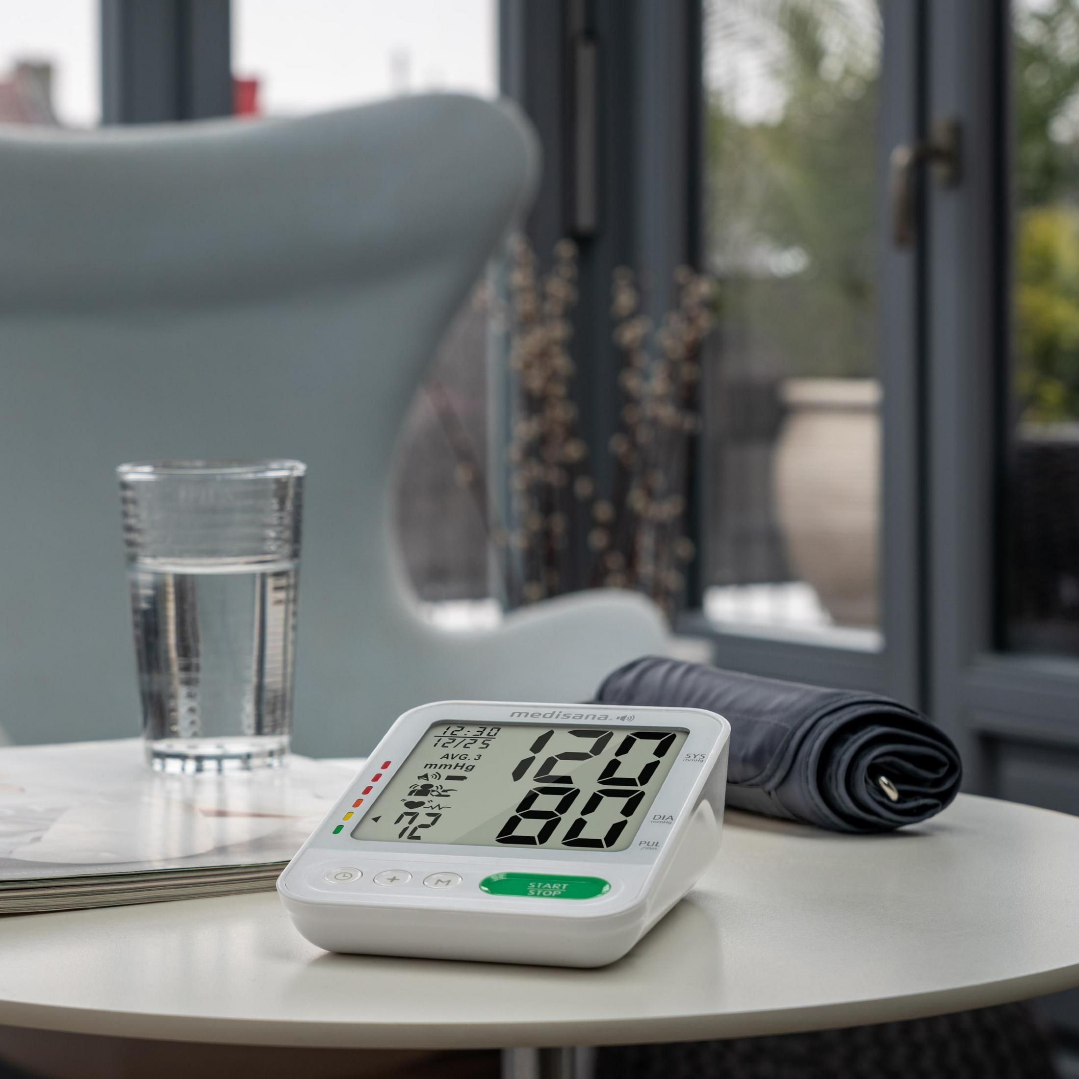 Cyfrowy monitor ciśnienia krwi, szklanka wody i zwinięty mankiet na stole.