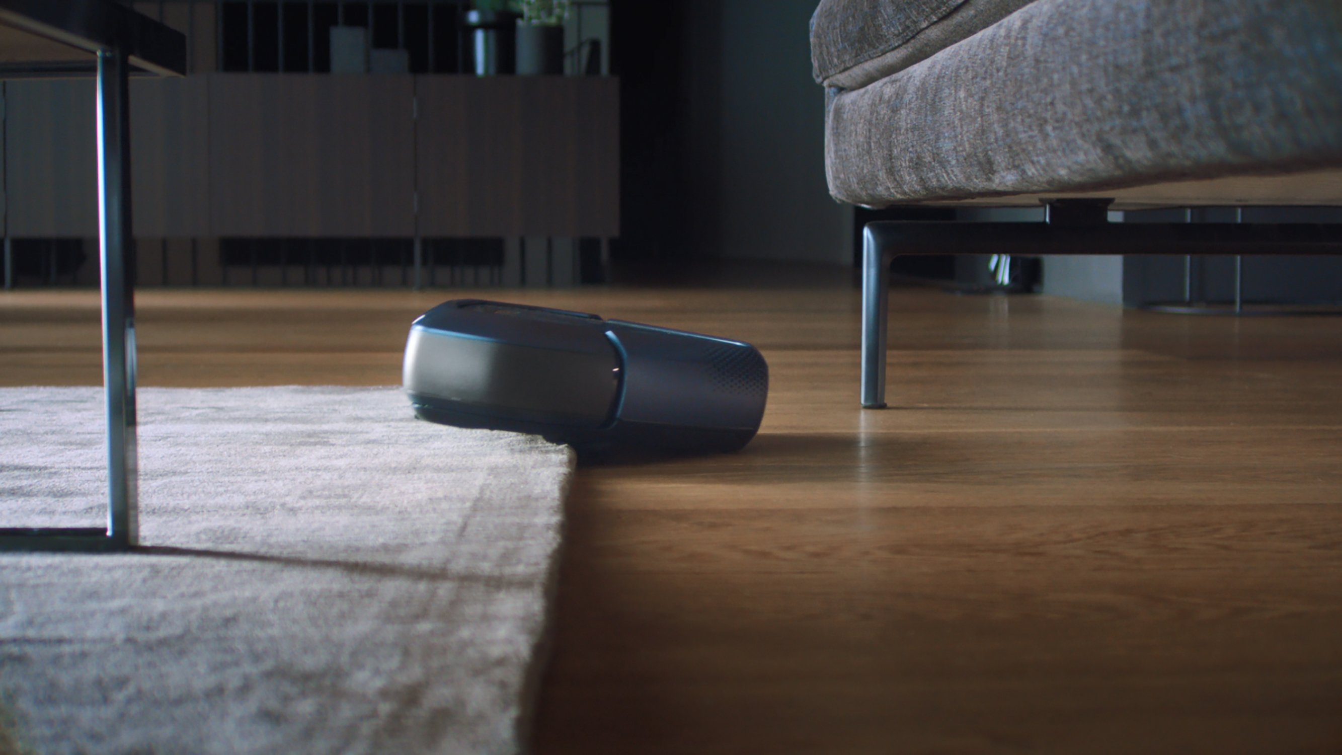 Robot sprzątający na podłodze z drewna, w pobliżu dywanu i sofy.