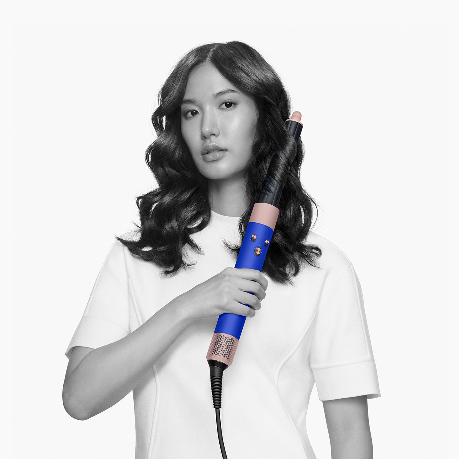 Donna che tiene in mano un arricciacapelli blu e rosa, con capelli scuri e che indossa una camicia bianca.