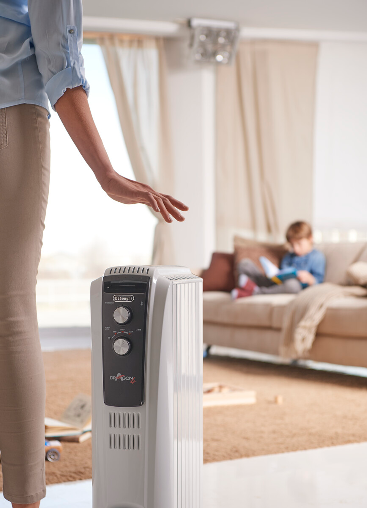 Dłoń kobiety w pobliżu białego grzejnika olejowego. Dziecko czyta na sofie w tle.