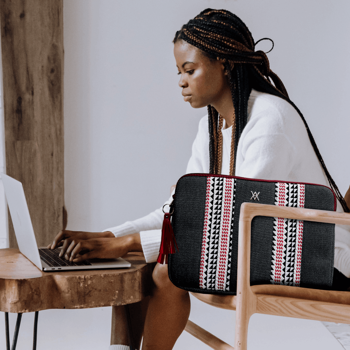 Mujer con trenzas usando una laptop. Bolso y funda con patrones en mesa. Silla de madera.
