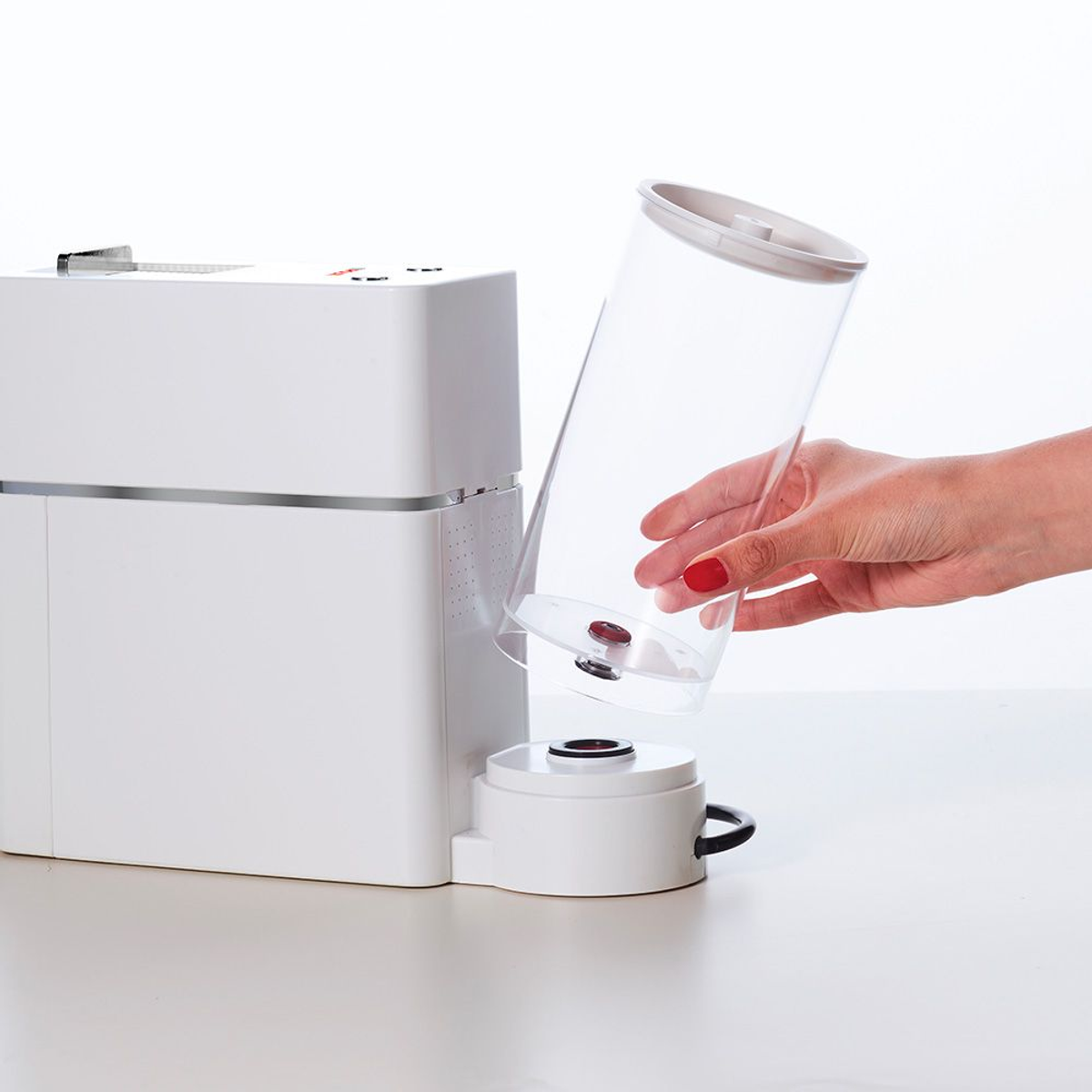 Una máquina blanca con un cilindro transparente sostenido por una mano, ambos sobre una superficie blanca.