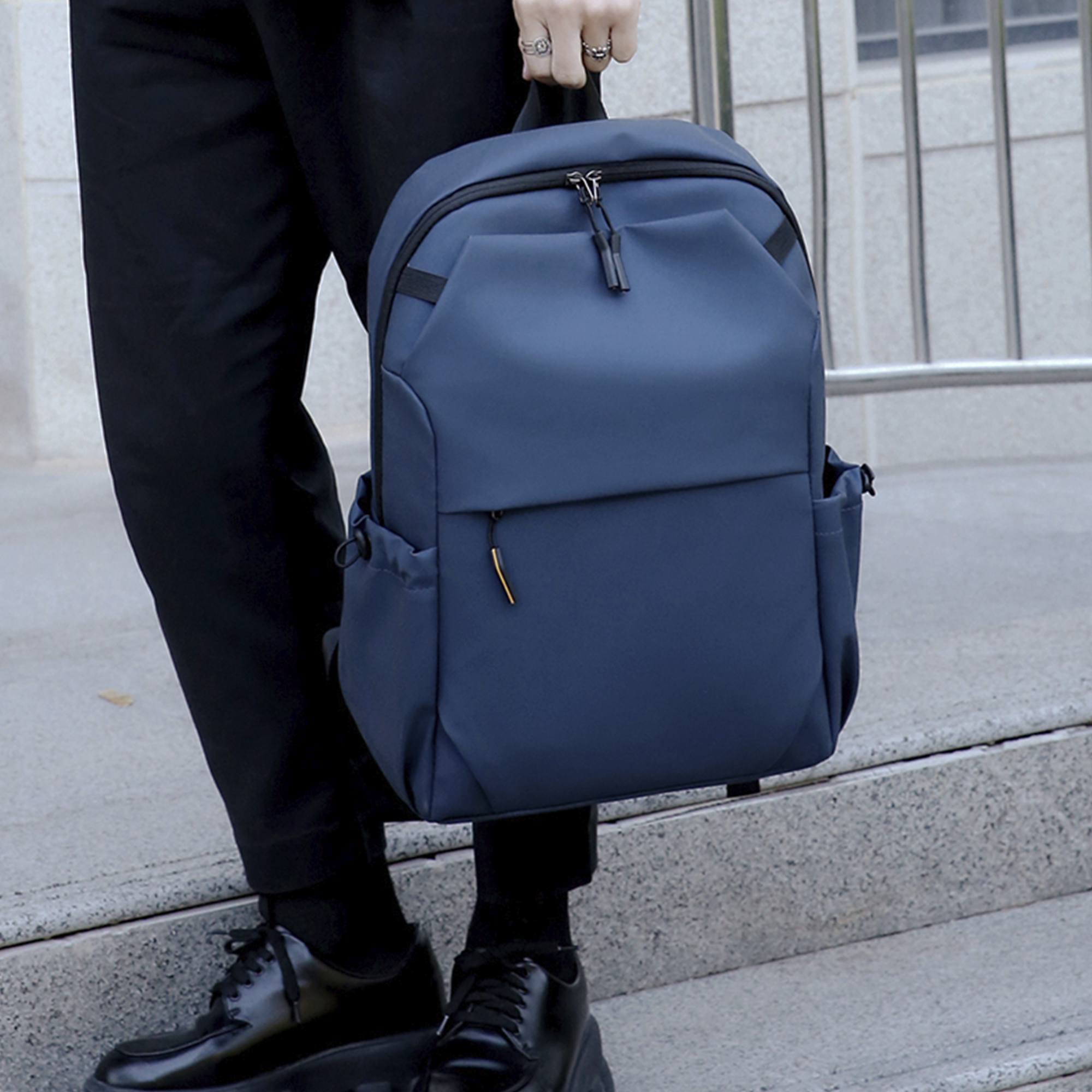 Persona che tiene uno zaino blu, in piedi sulle scale, con pantaloni e scarpe nere.