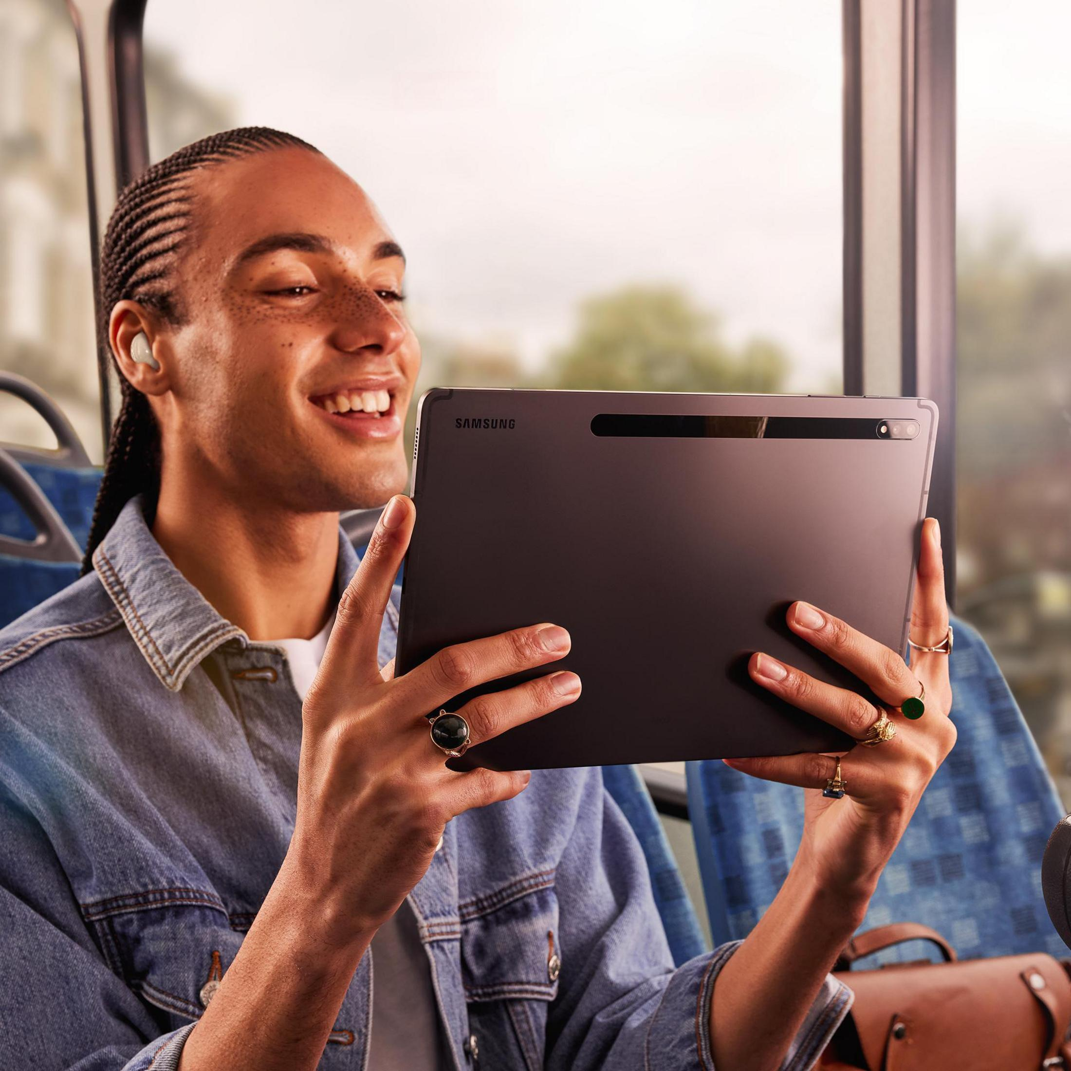 Mężczyzna trzymający tablet. Uśmiecha się i ma na sobie dżinsową kurtkę. Ma kolczyki i kilka pierścieni.