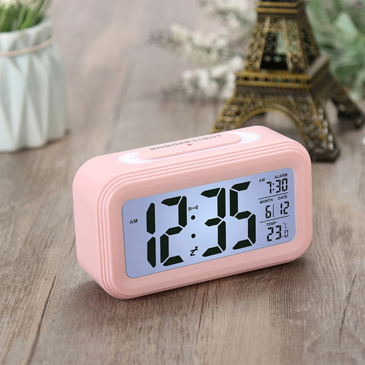 Pinkfarbener digitaler Wecker mit Uhrzeit und Datum, kleiner Vase und Turm im Hintergrund.