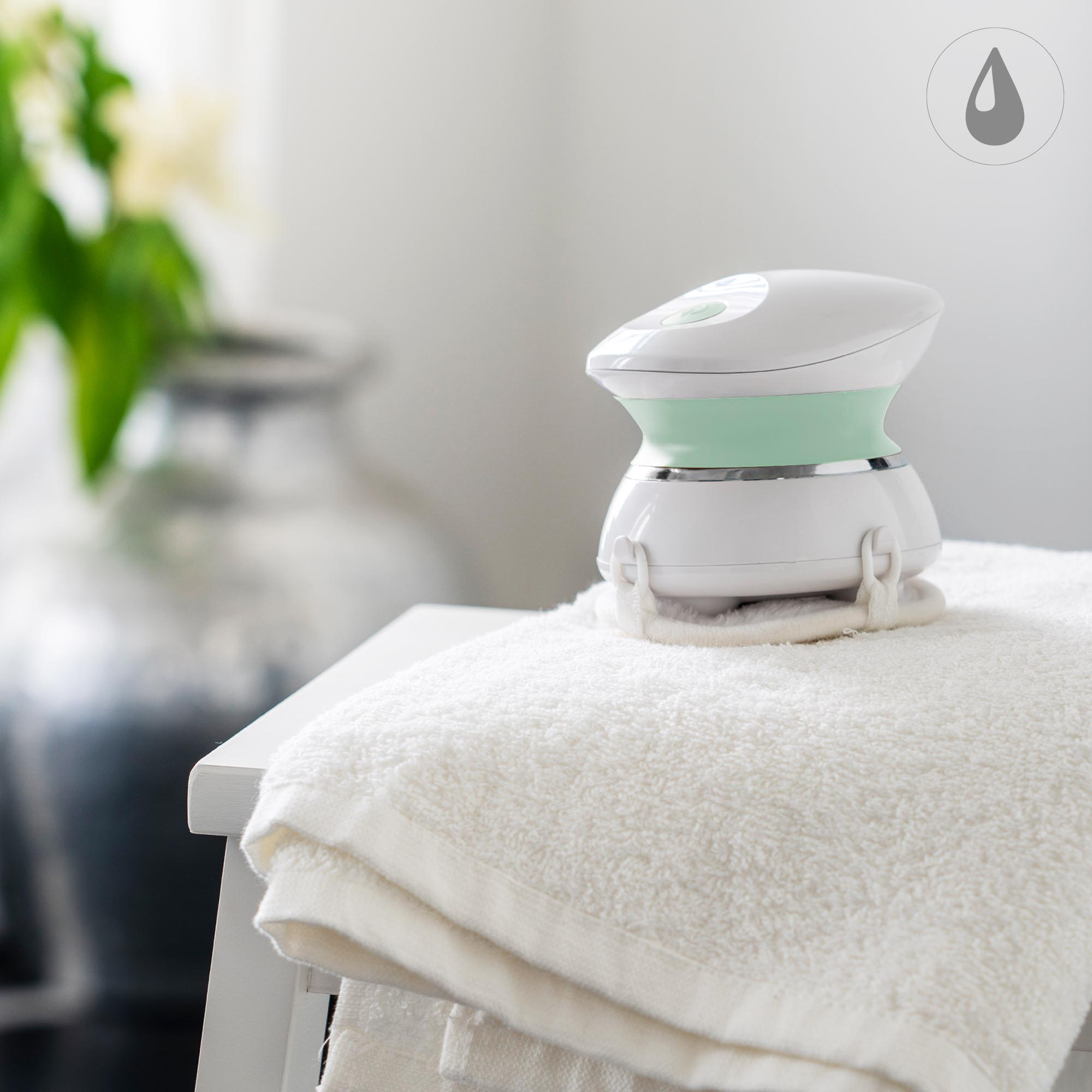 Un dispositivo di bellezza bianco e verde è su un asciugamano bianco su uno sgabello bianco, con un'icona a goccia grigia.
