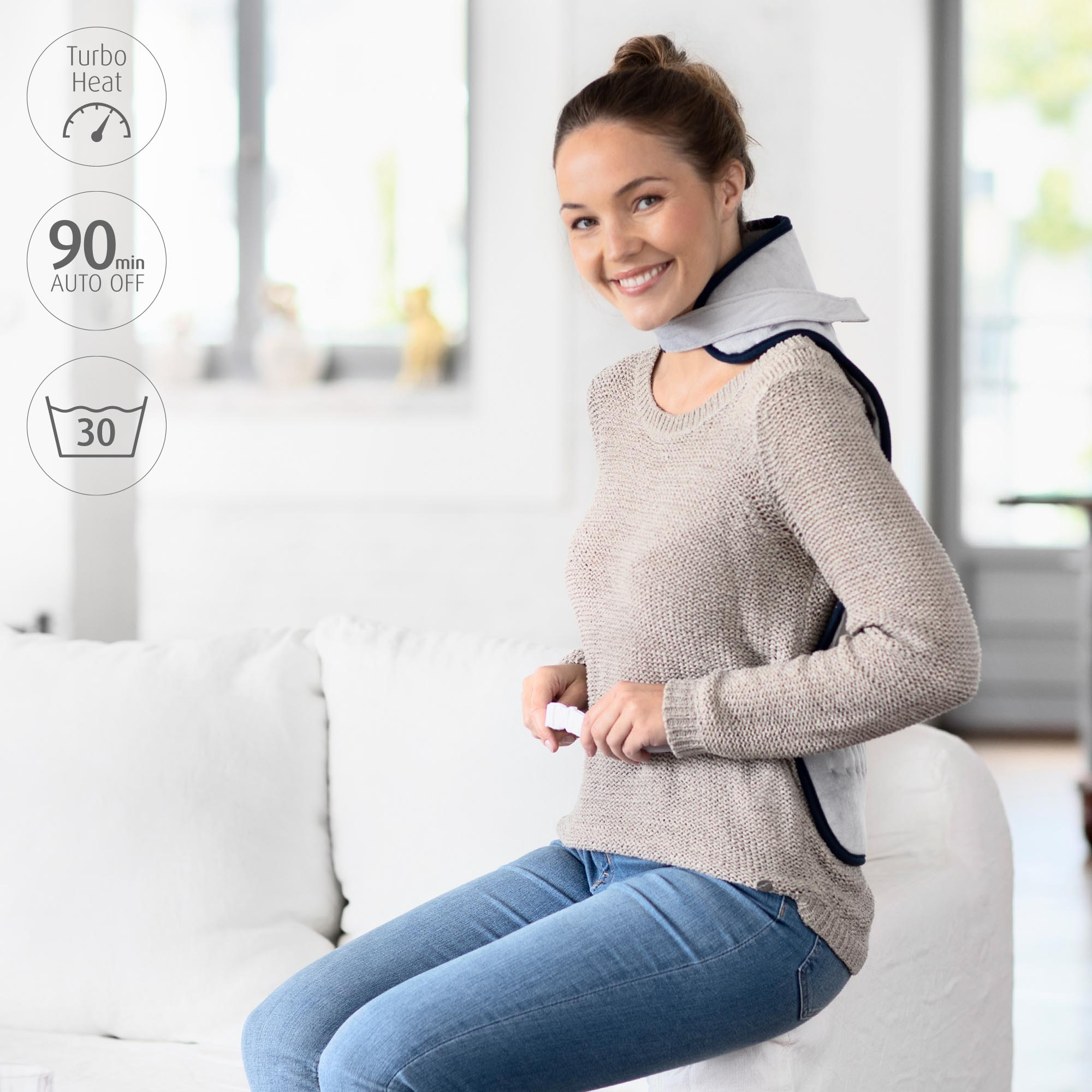 Kobieta siedzi na kanapie, ma poduszkę rozgrzewającą szyję i uśmiecha się do kamery.