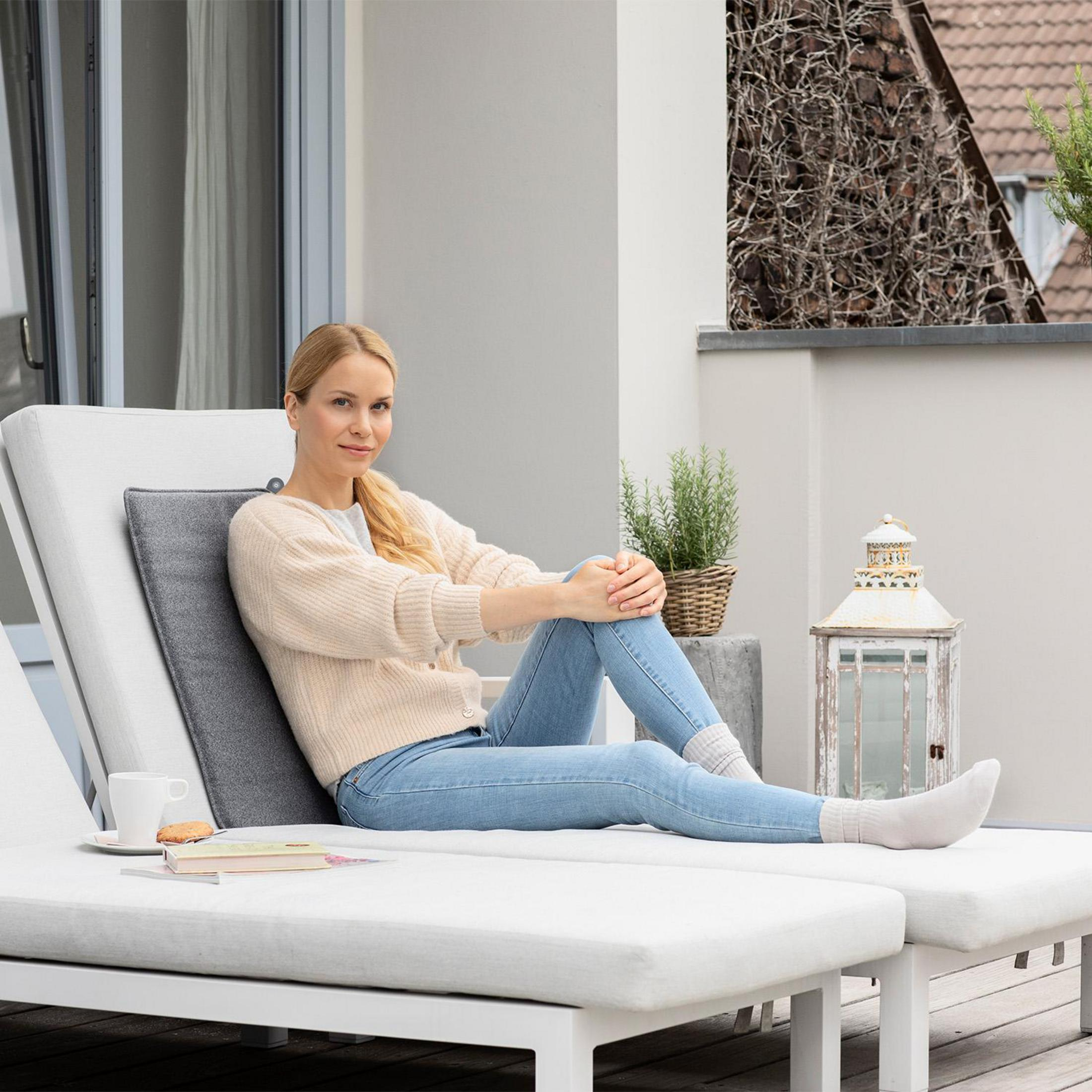 Kobieta siedzi na białym leżaku. Tło tarasu. Książka i filiżanka na stole.