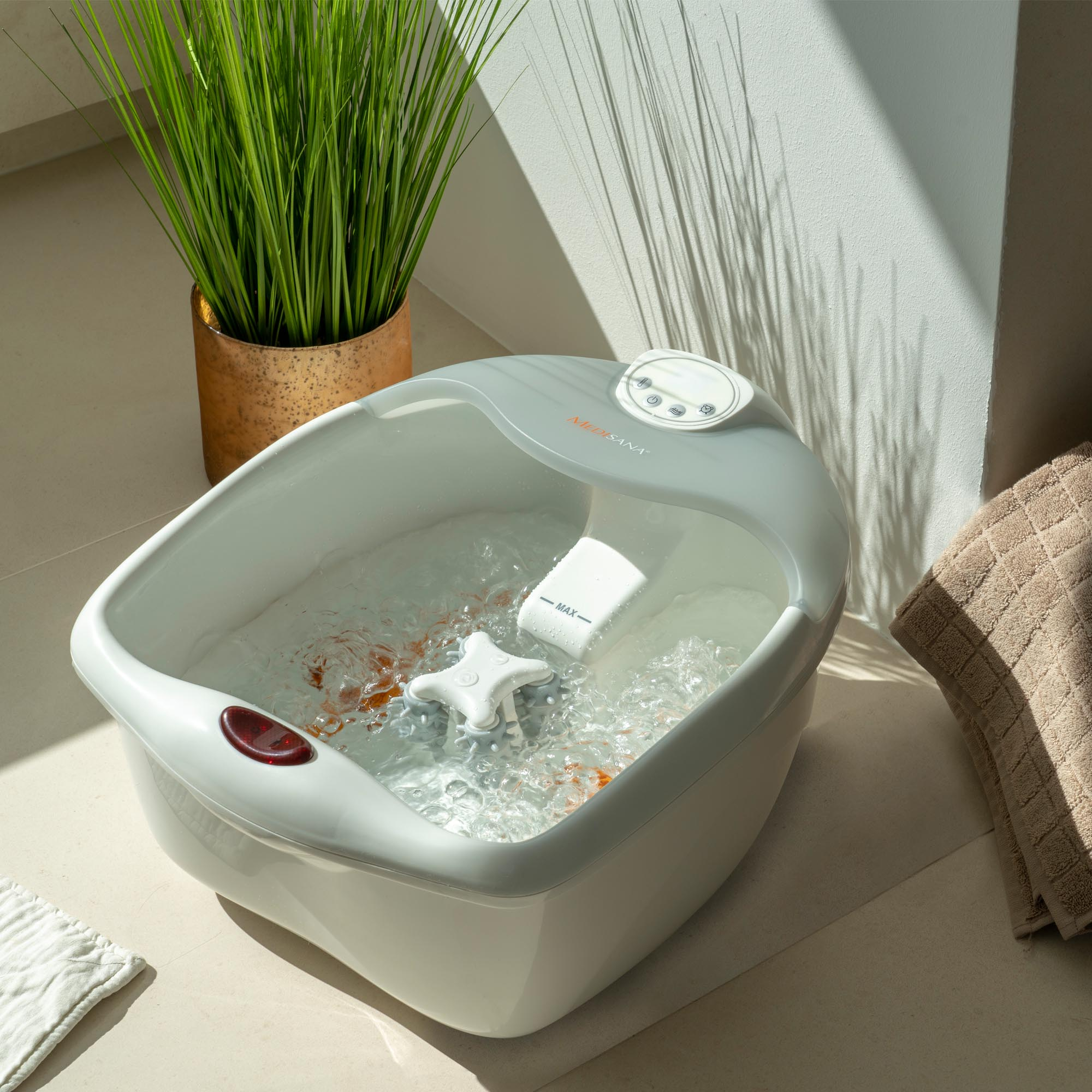 Foot spa with water and pebbles. Green plant in a copper pot on the left. Towel on the right.