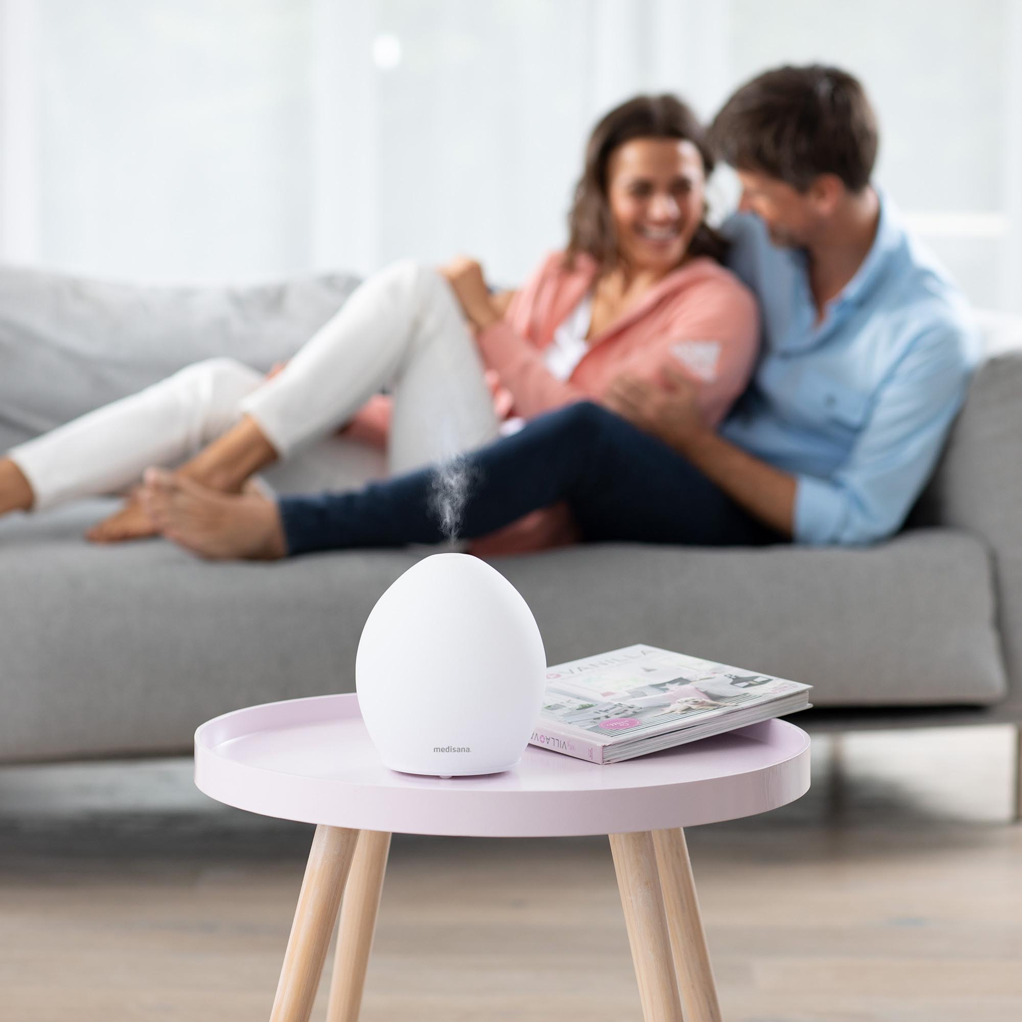 A white aroma diffuser on a pink table. A couple is on a gray couch.