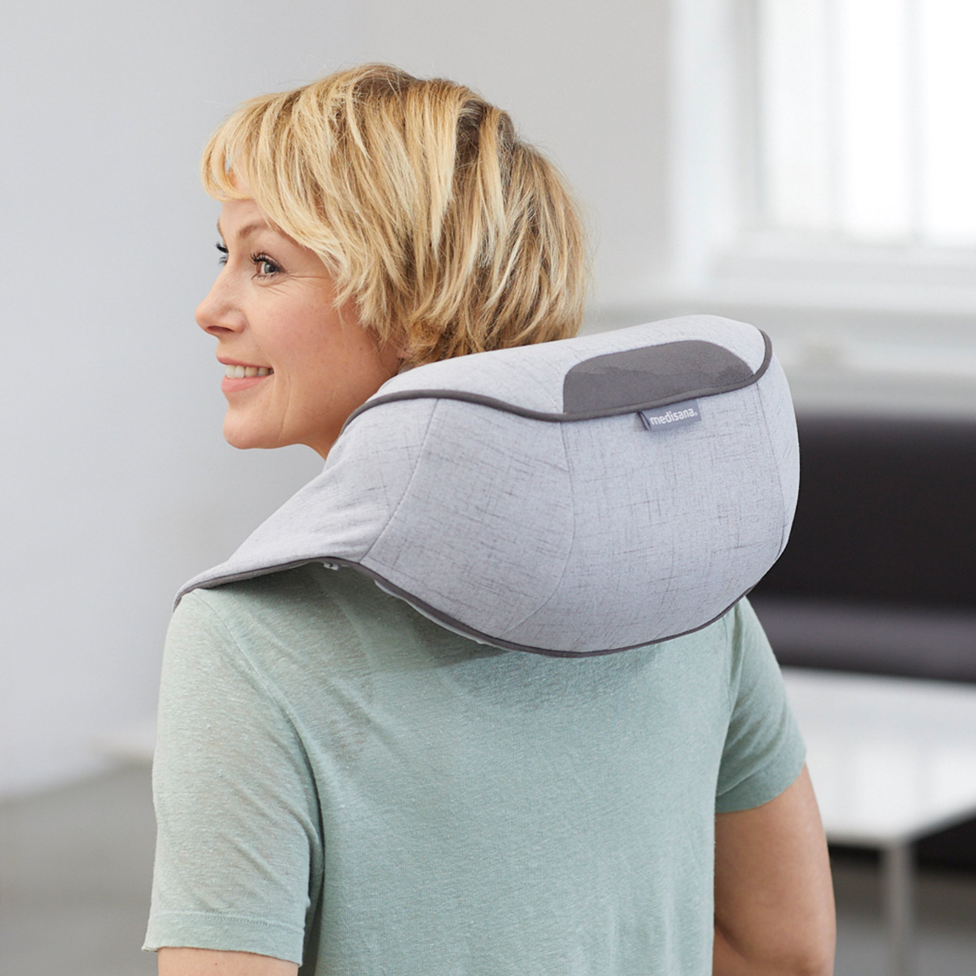 Una mujer rubia sonriendo mientras usa un masajeador de cuello gris.