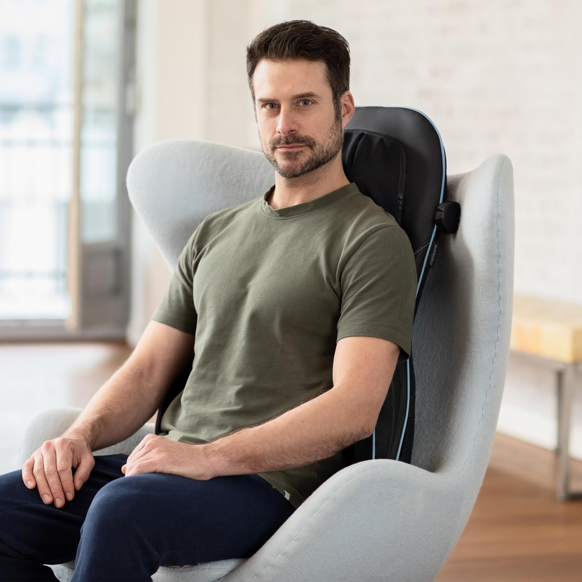 Mężczyzna siedzi w szarym fotelu, z czarną poduszką do masażu na plecach. Ma na sobie zieloną koszulę.