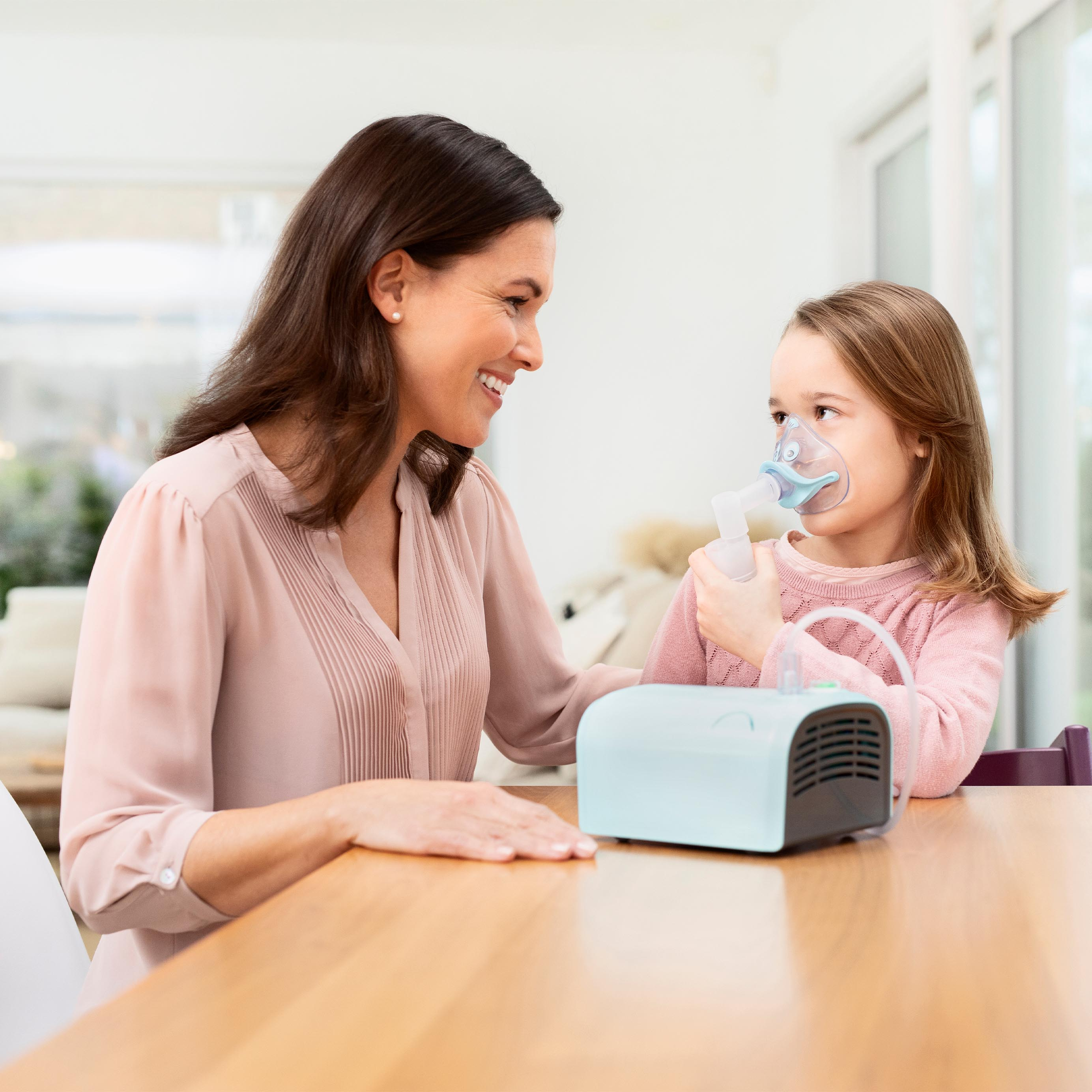 A woman and a girl using a nebulizer. They are sitting at a wooden table indoors.