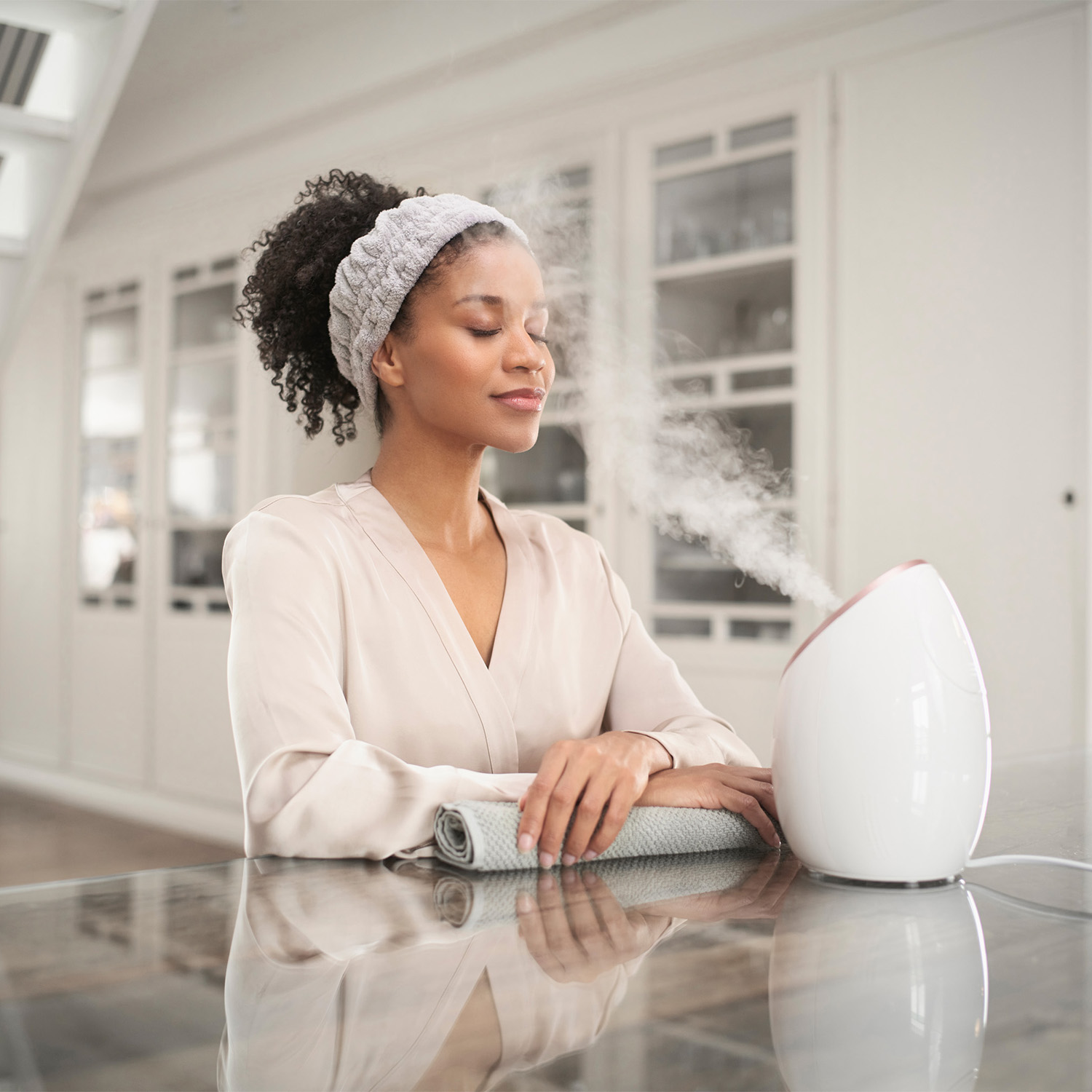 Femme utilisant un steamer facial, vapeur montante. Elle porte un bandeau, assise à une table.