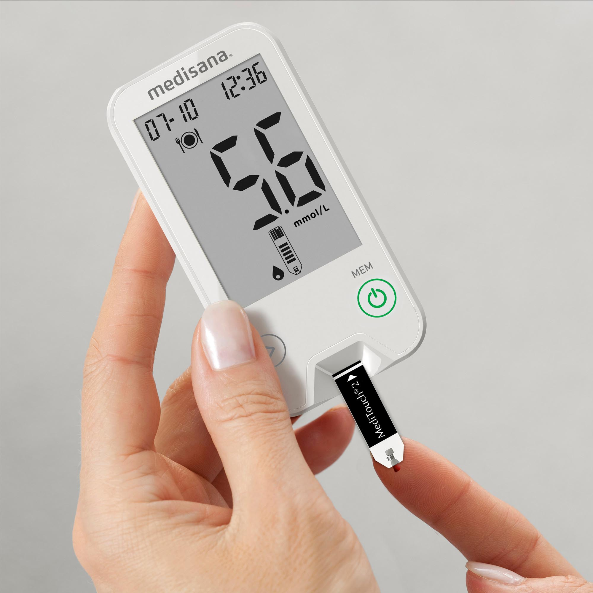 Hand holding a white Medisana blood glucose meter with a test strip inserted and a reading of 5.6.