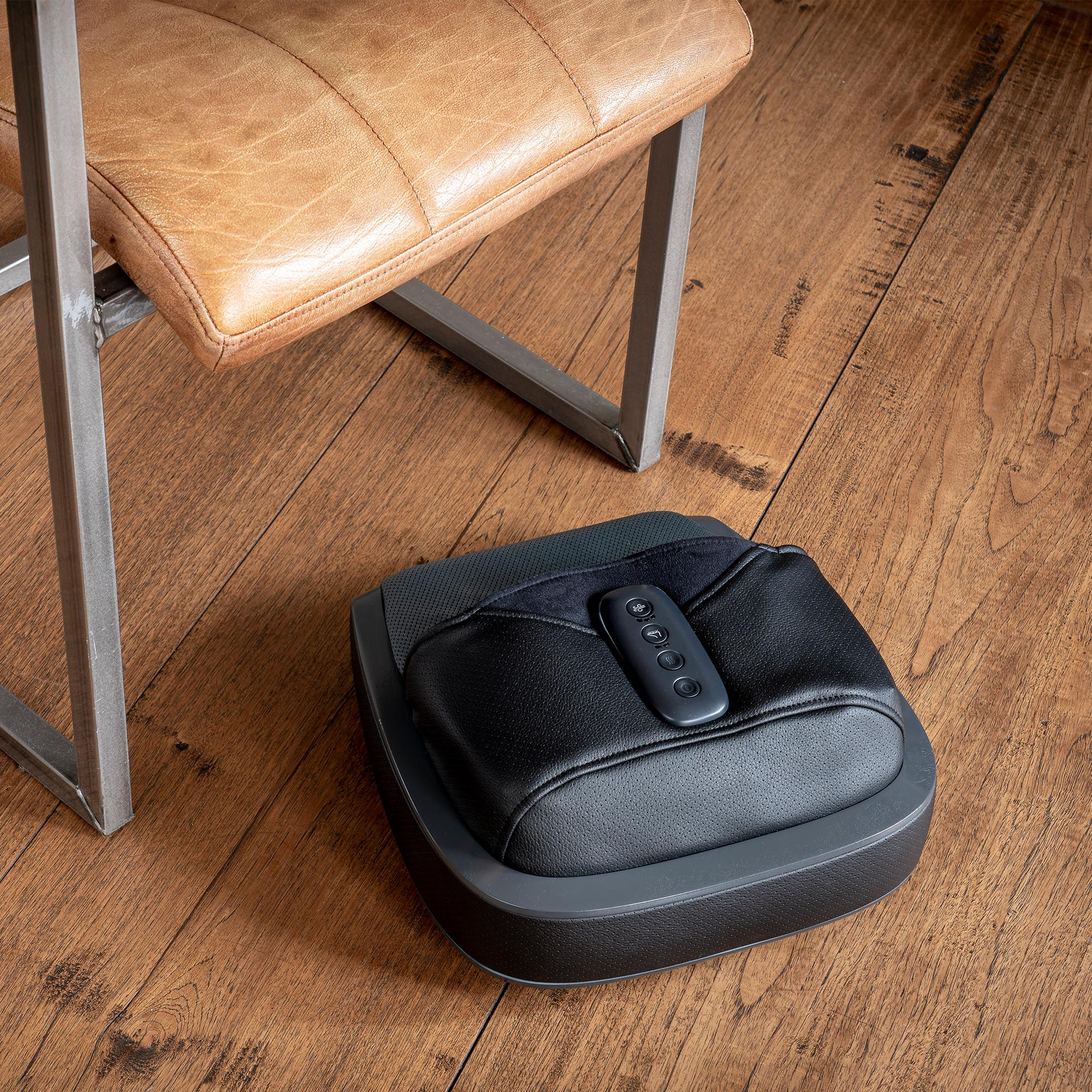 Masażer do stóp z pilotem umieszczony na drewnianej podłodze. Z lewej strony skórzany fotel.