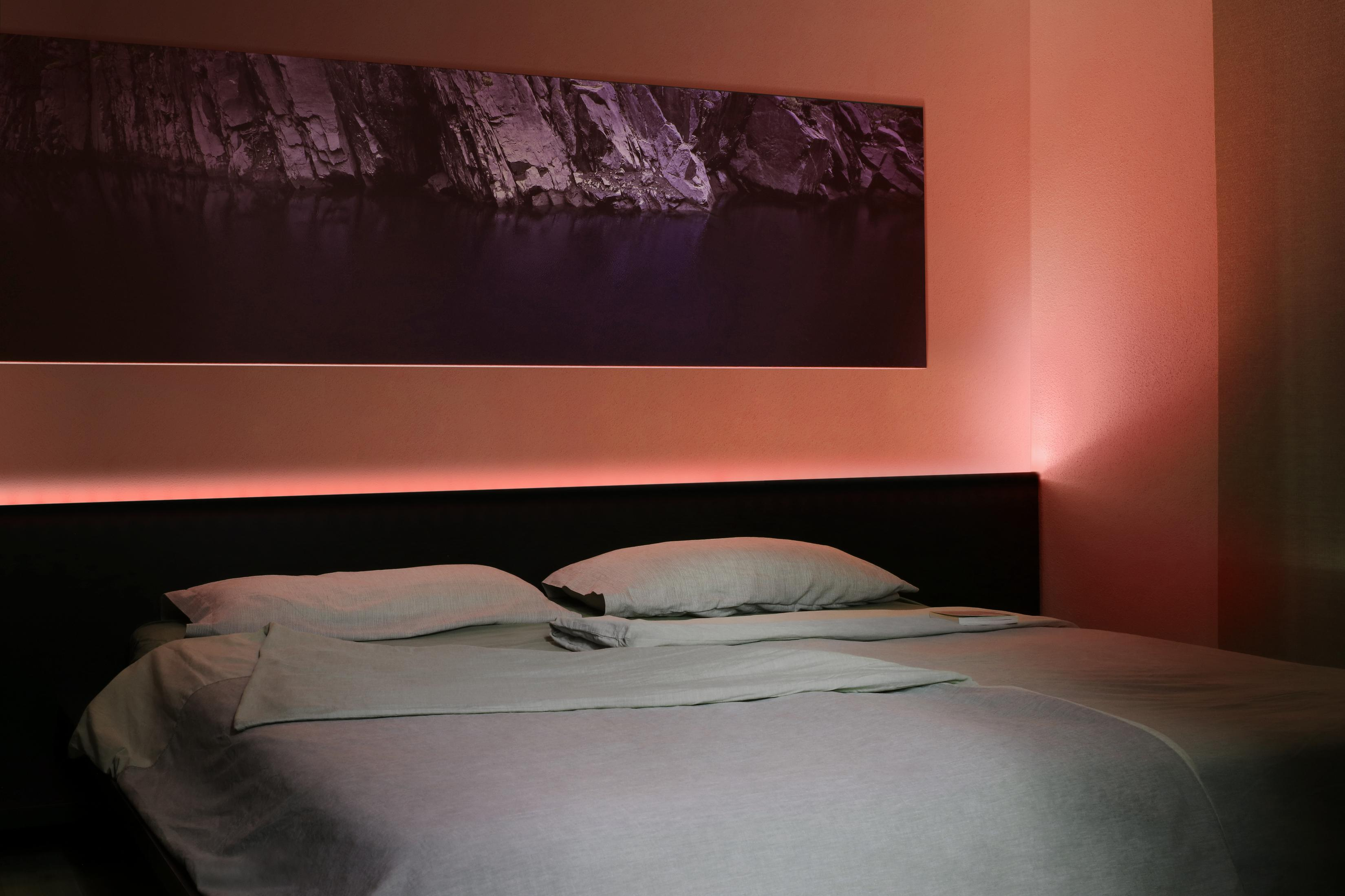 Scena della camera da letto. Letto con cuscini e coperta, illuminato da luci rosse sopra. Immagine sul muro.