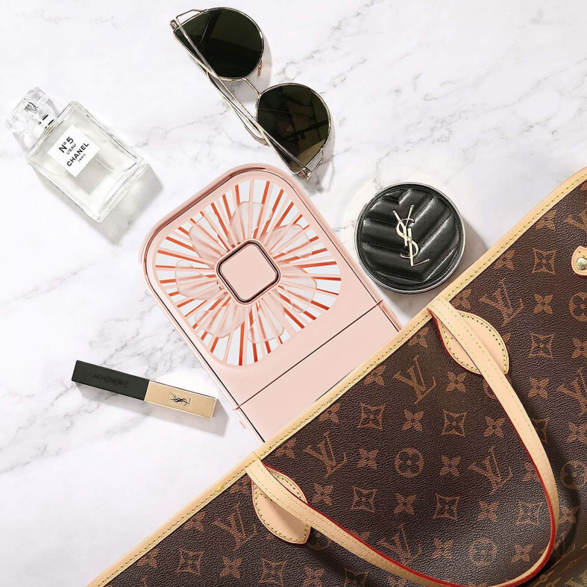 Ein Flatlay einer Parfümflasche, Sonnenbrille, einem kleinen Ventilator, einem Make-up-Compact und einer Louis Vuitton Tasche.