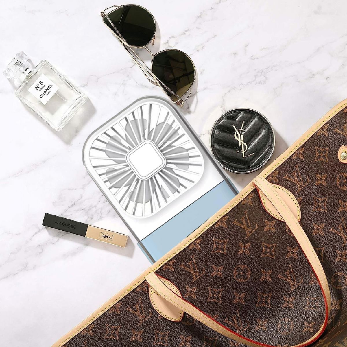Flach liegend: Handtasche, Parfüm, Sonnenbrille, tragbarer Ventilator und Make-up auf Marmor.