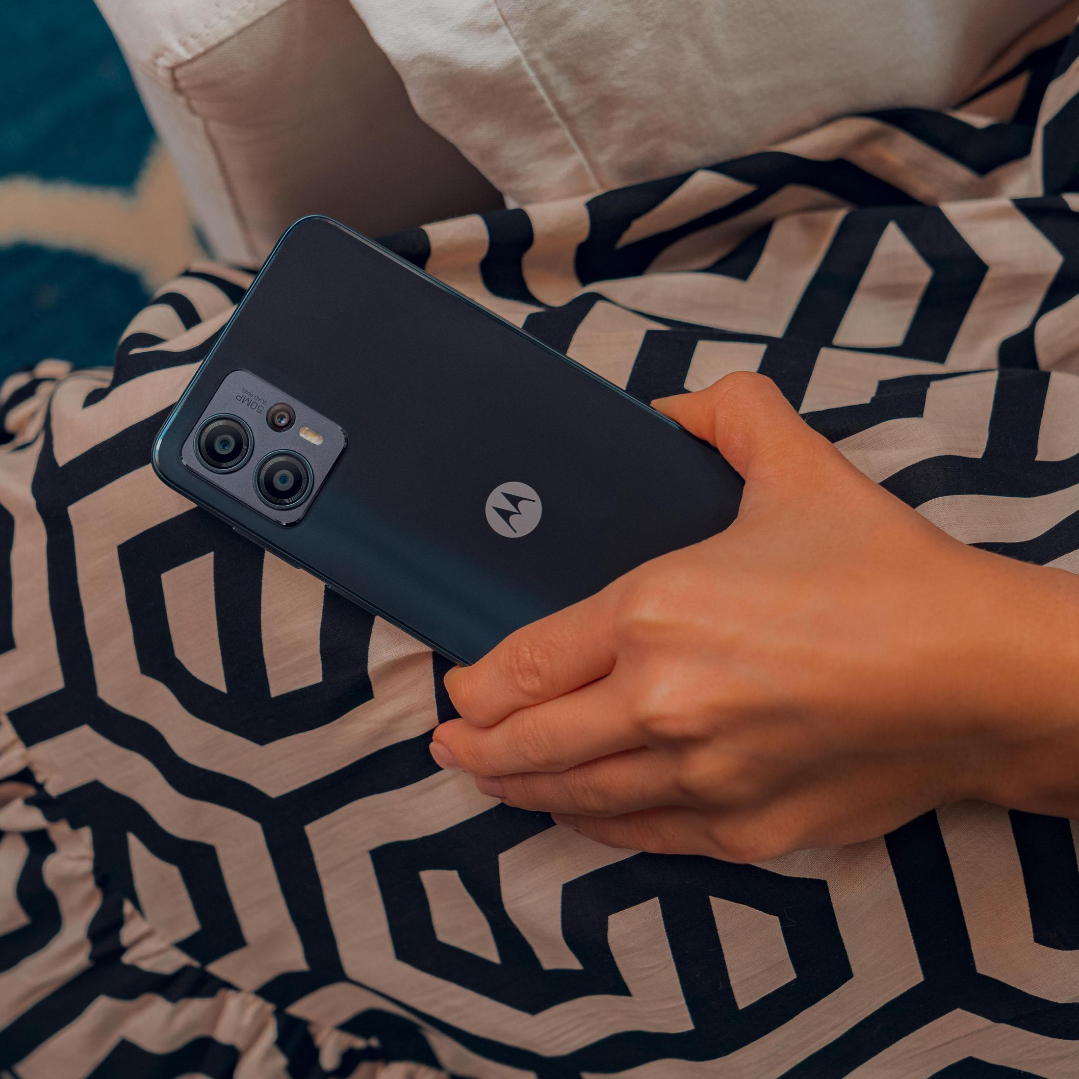 Osoba trzymająca czarny smartfon z dwoma widocznymi aparatami na wzorzystej tkaninie.