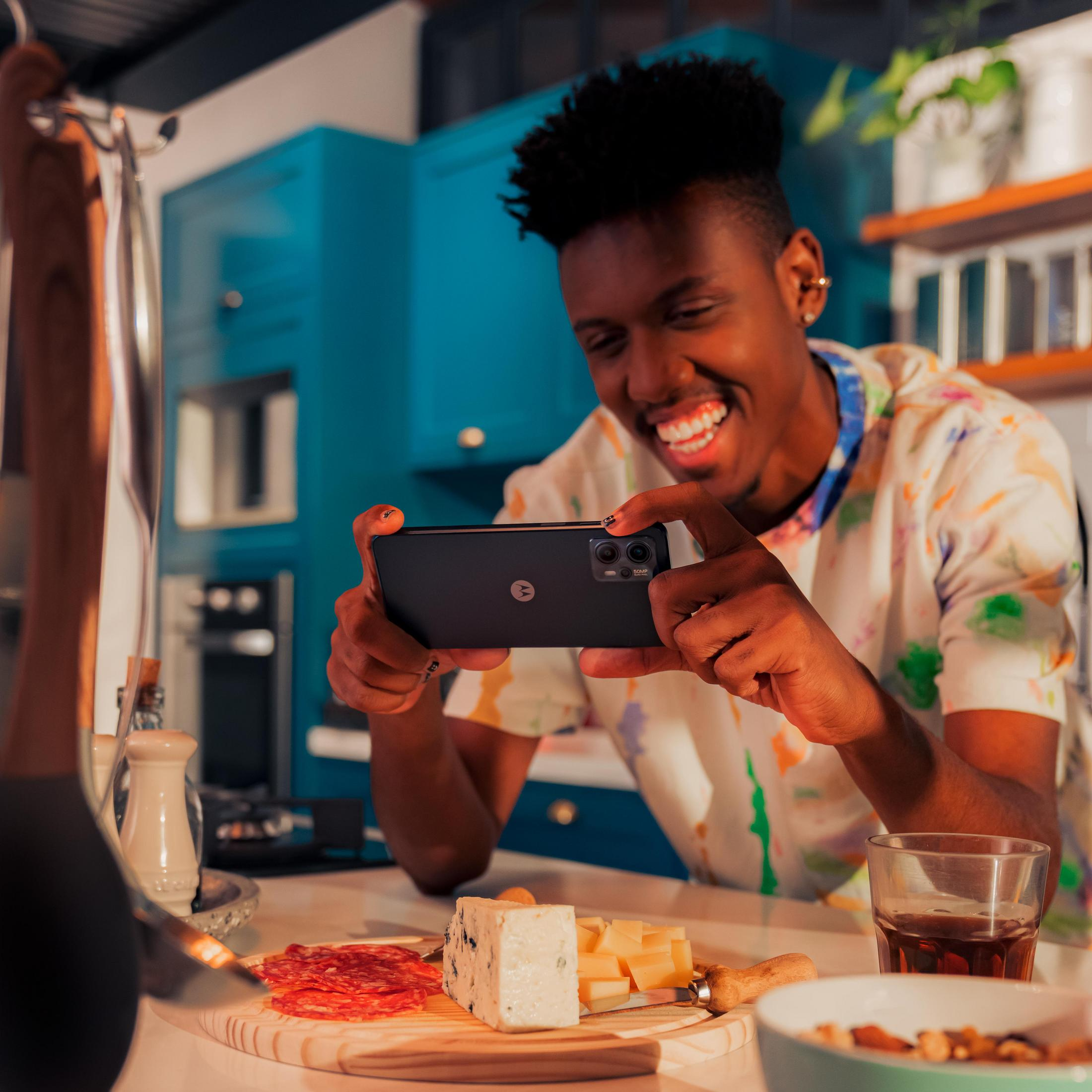 Uśmiechnięty mężczyzna robi zdjęcie jedzenia na drewnianej desce telefonem. Tło kuchni z niebieskimi szafkami.