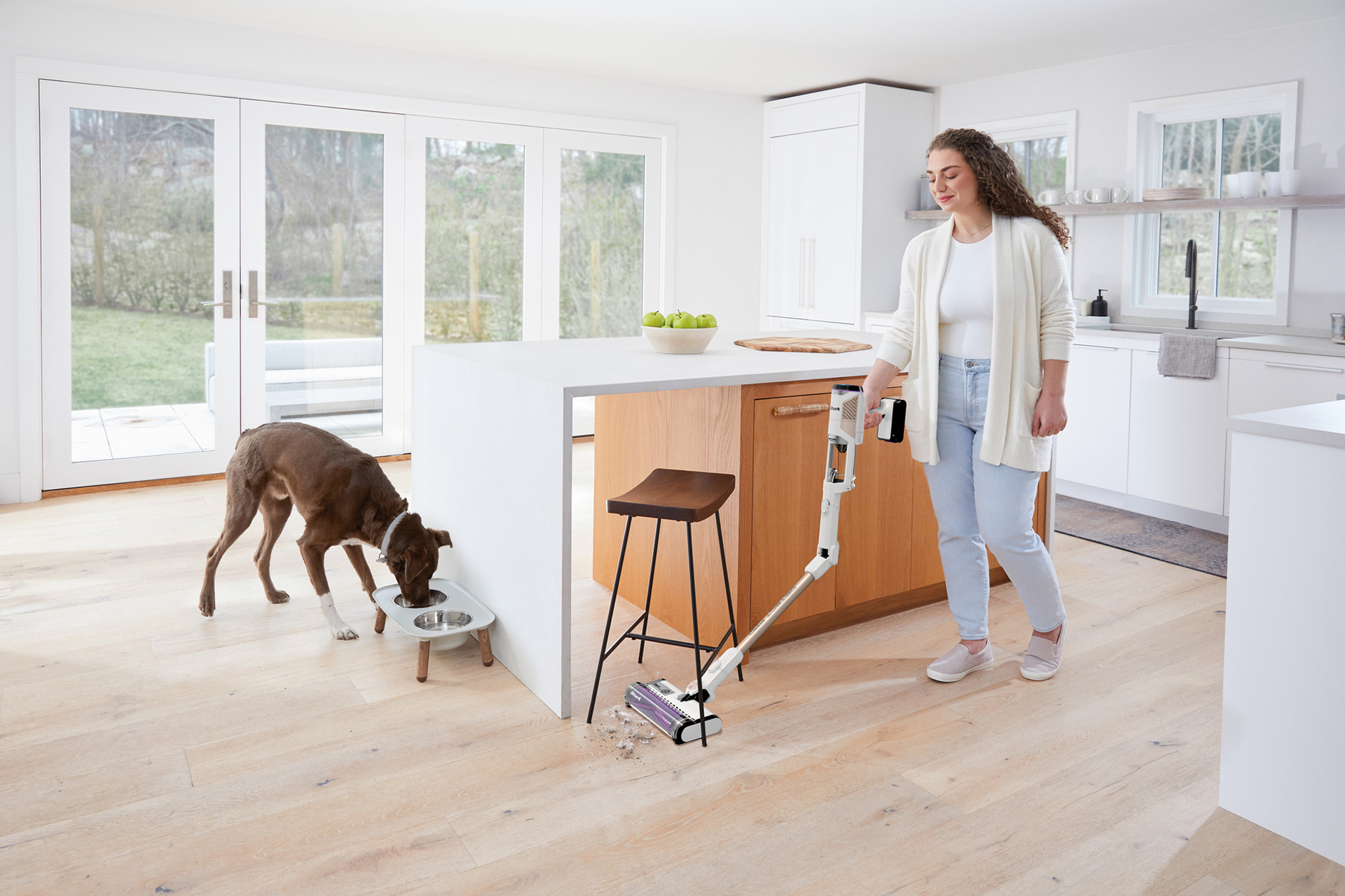 Mujer aspira el suelo de la cocina. Un perro come de un cuenco. Se ven armarios de madera y superficies blancas.