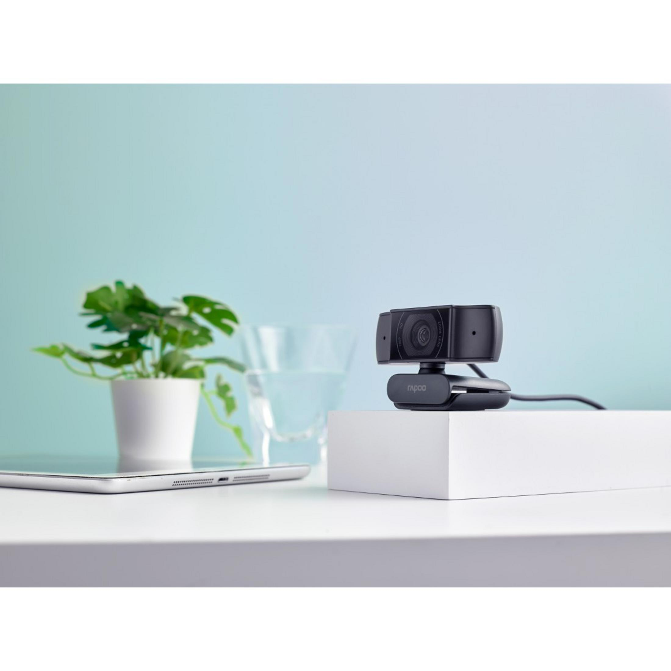 Kamera internetowa na białym stojaku z rośliną, szklanką wody i tabletem. Jasnoniebieskie tło.
