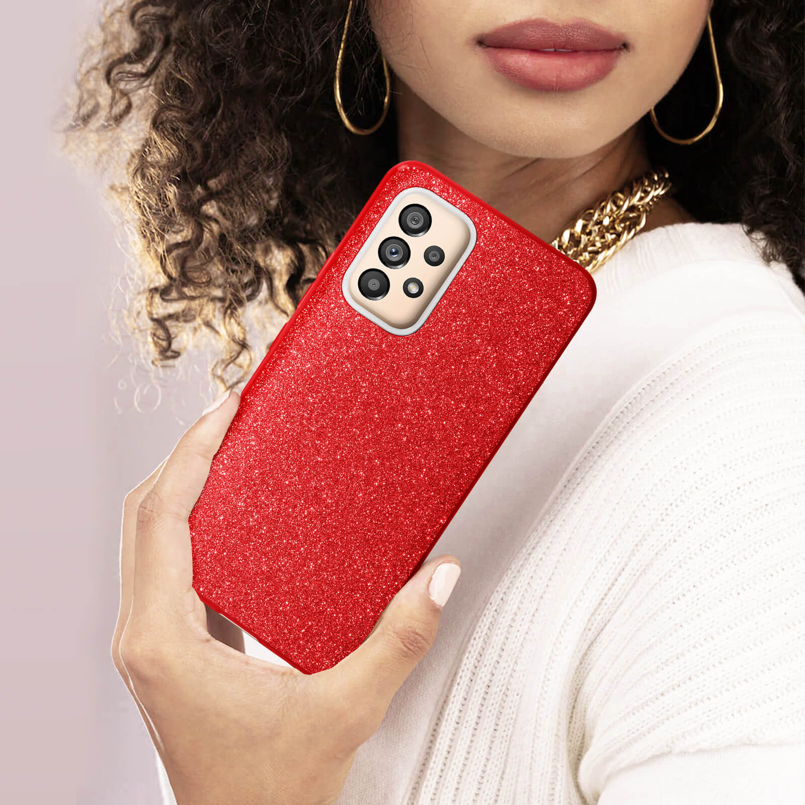 Une femme tenant une coque de téléphone rouge pailletée. Elle porte un pull blanc et des bijoux dorés.