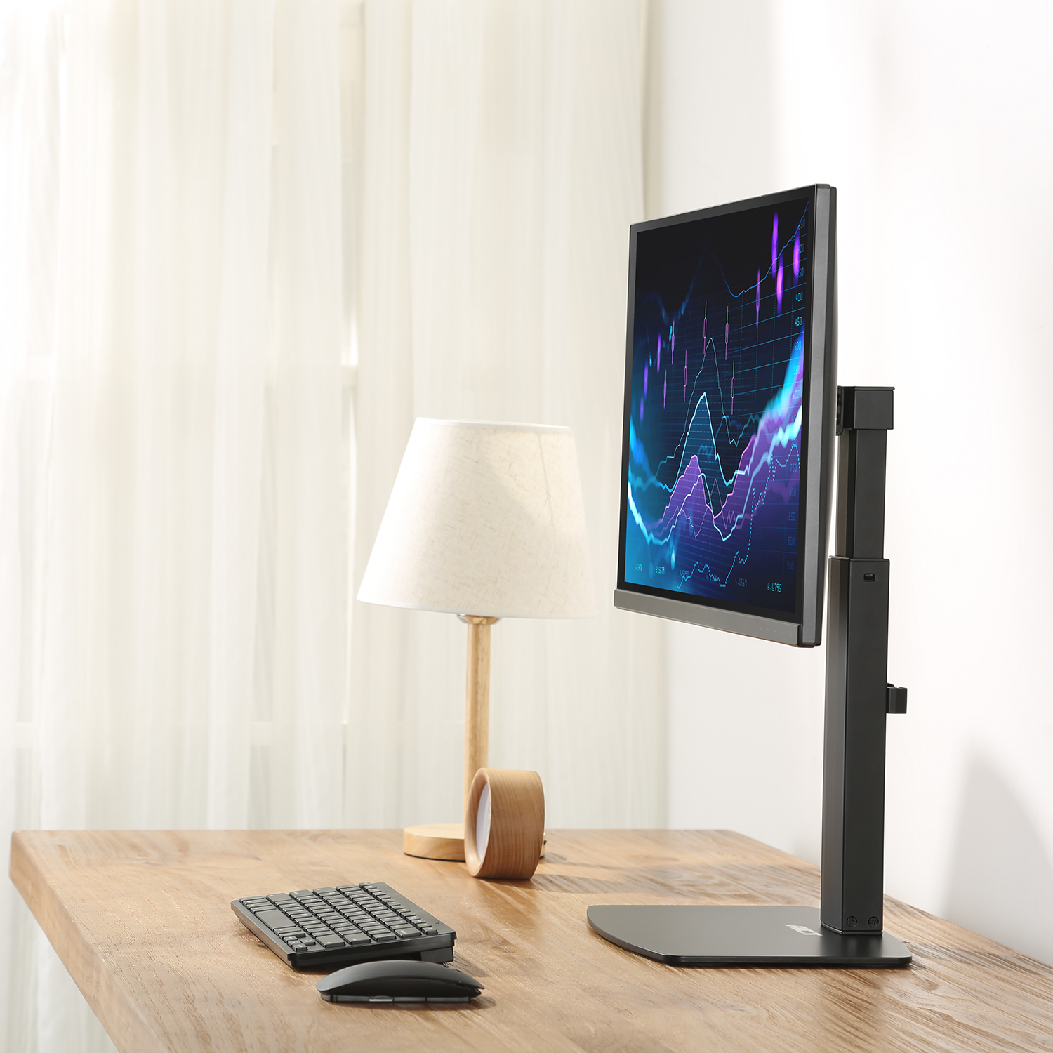 Een monitor op een standaard, een lamp, toetsenbord en muis op een houten tafel, een wit gordijn op de achtergrond.