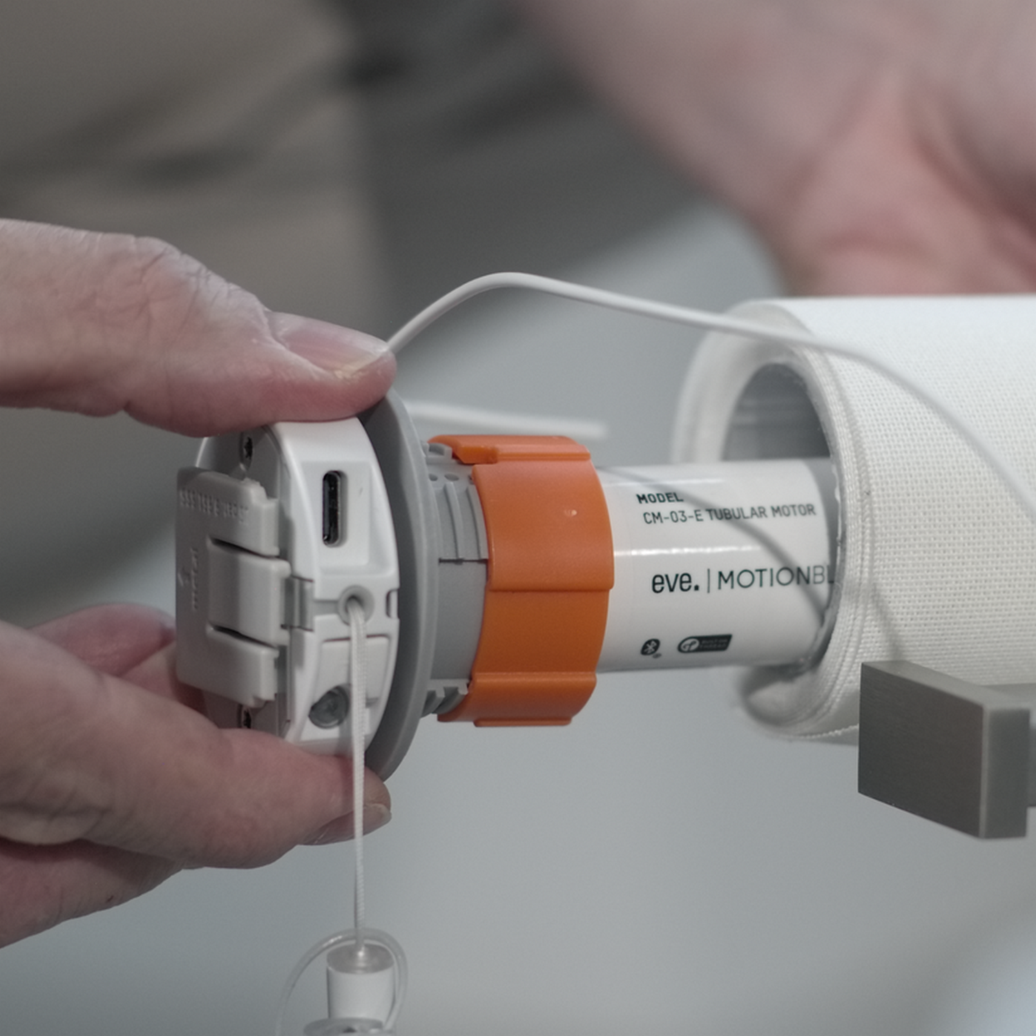 Mano che tiene un dispositivo bianco e arancione collegato a una tenda a rullo bianca. Il dispositivo è etichettato 'EVE MOTIONBLINDS'.