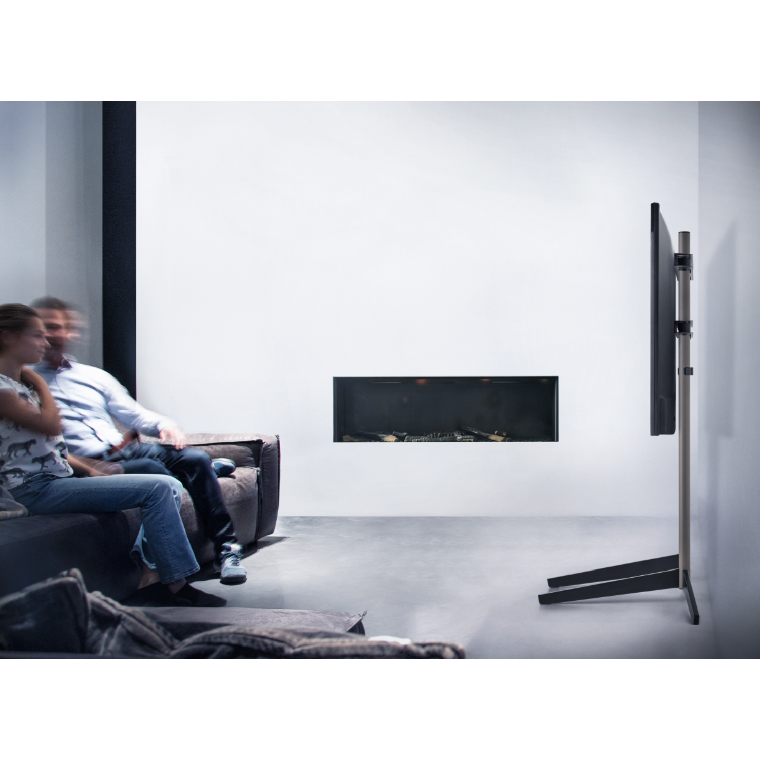Un couple assis sur un canapé regarde une télévision montée sur un pied. Fond de mur blanc.