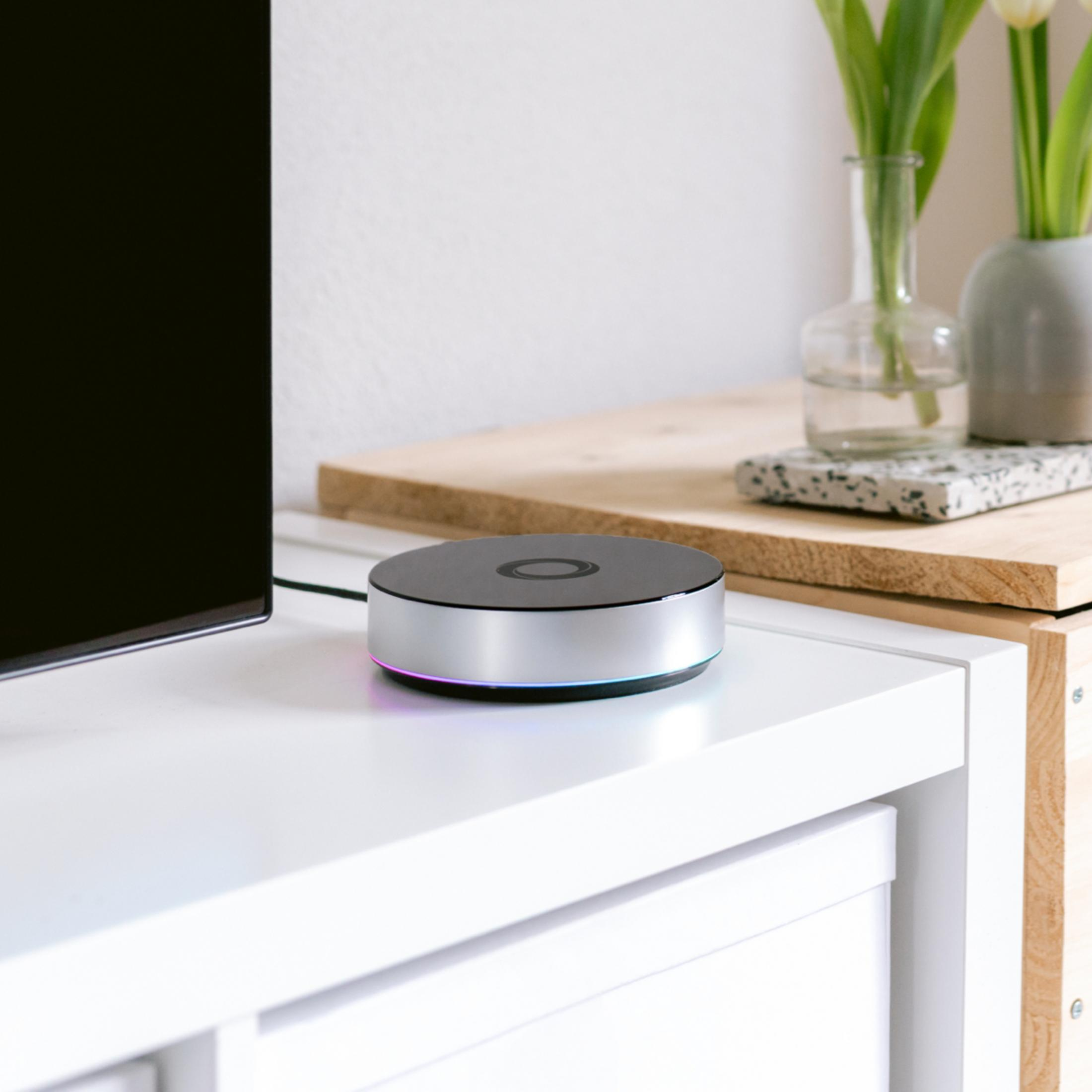 Schwarzes und silbernes Smart-Home-Gerät auf weißer Oberfläche. Hintergrund mit Pflanzen und TV.