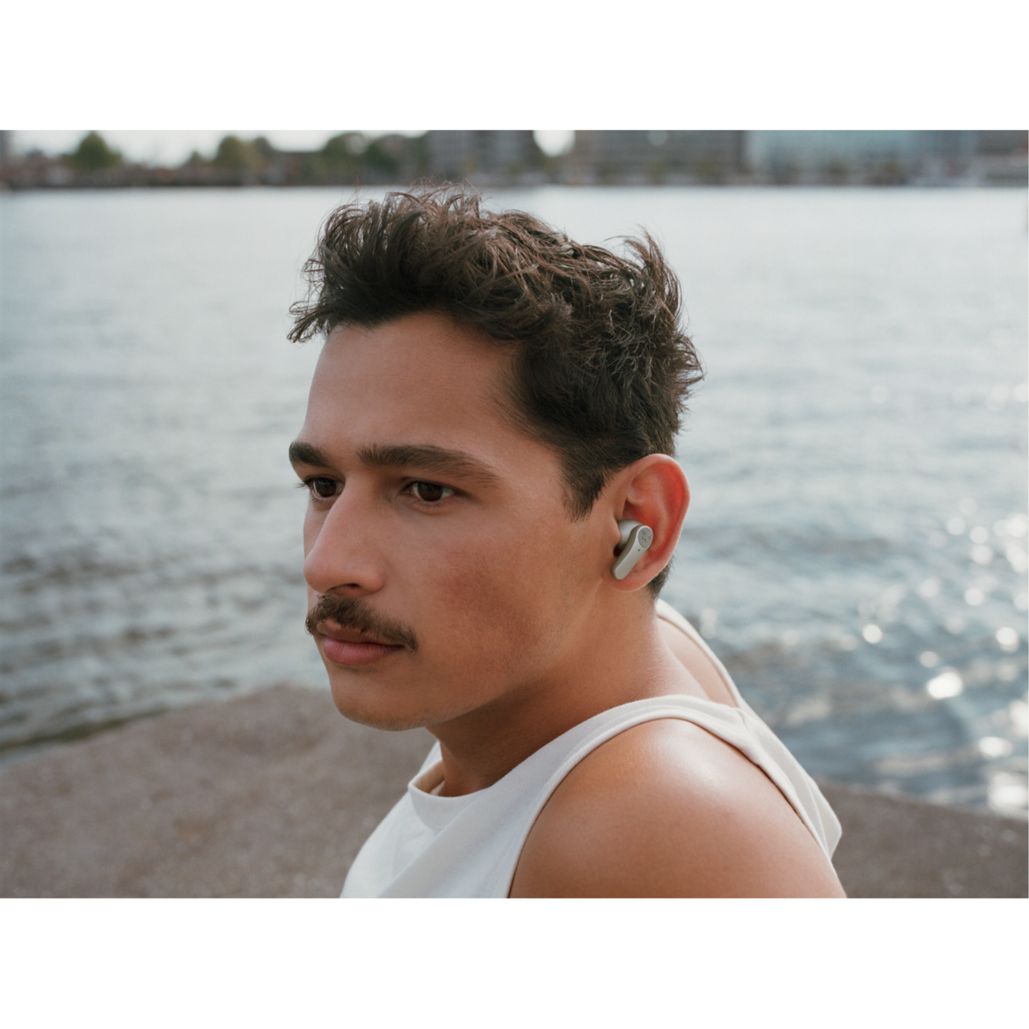 Mężczyzna z wąsami w białym podkoszulku, noszący szare słuchawki. Tłem jest woda.